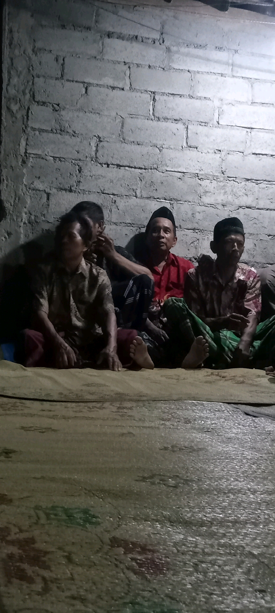 Four men are seated on a woven mat against a rough, light-colored brick wall. The man on the far left is mostly obscured but appears to be wearing a patterned shirt and dark pants. Next to him, a man in a light brown, patterned shirt is looking down and to his left, with one hand resting on his knee and the other near his face. He is wearing maroon pants. To his right, a man in a red shirt and a black peci hat is looking forward. The man on the far right is also wearing a peci hat and a patterned shirt with red and white elements, and is looking forward. He is wearing green and white checkered pants. The floor is covered with a light brown mat that has some faded colorful patterns. The lighting is dim, casting shadows on the wall. The scene appears to be indoors, with a rough, unfinished wall suggesting a utilitarian or less formal setting. Based on the context of Kedungmulyo, Indonesia, this could be a gathering in a home or community space.