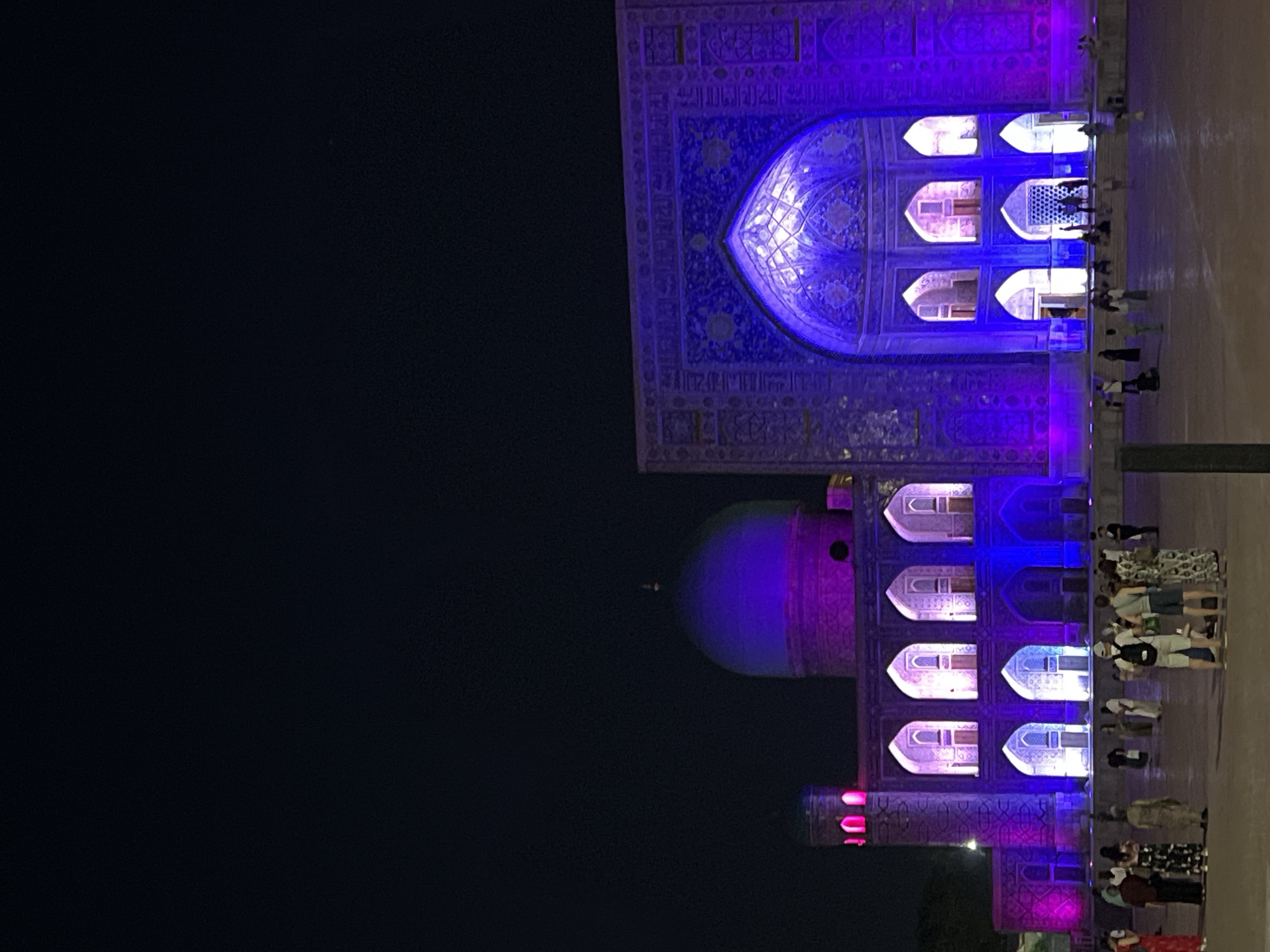 The image depicts a nighttime view of the Registan Square in Samarkand, Uzbekistan. The Gur-e-Amir mausoleum and other structures are illuminated with vibrant purple and pink lights, highlighting their intricate architectural details and mosaic tilework. A group of people are visible in the foreground, some standing and observing the illuminated buildings, while others are walking or conversing. The sky is dark, suggesting it is late evening or night. The ground is paved, and there are no other significant environmental features visible beyond the square itself. The lighting creates a dramatic and colorful atmosphere, emphasizing the grandeur of the historical site.