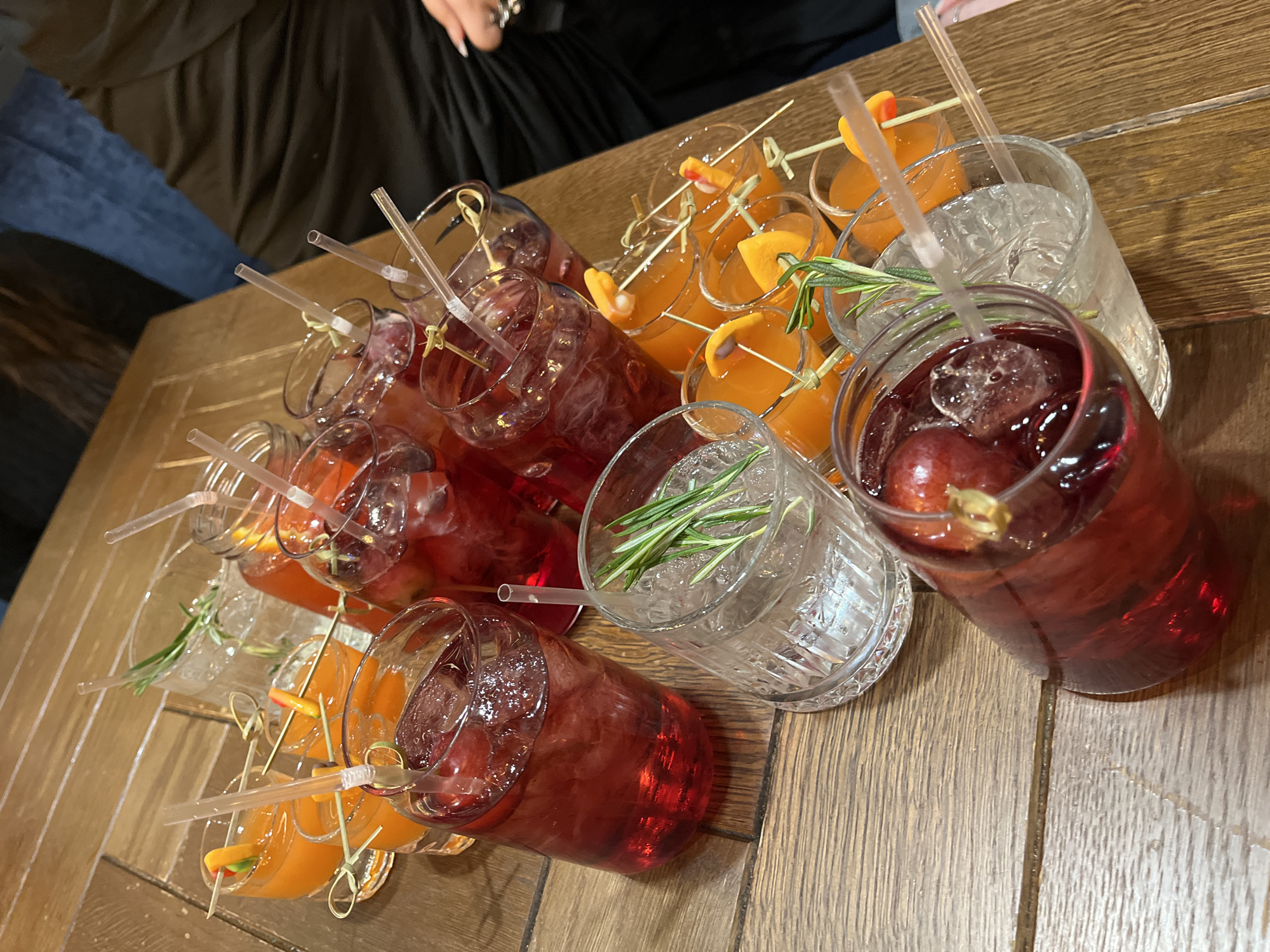 An overhead shot captures an array of cocktails arranged on a wooden table. The table features light brown wooden planks with visible grain, running diagonally across the frame.
The majority of the image is filled with glasses containing different beverages. There are at least three distinct types of drinks:
1. **Reddish-purple drinks:** These are prominent, filled with ice and garnished with grapes. They appear in several glasses, some with clear straws.
2. **Orange-colored drinks:** Several glasses contain a vibrant orange liquid, also with ice. These are garnished with small orange slices, often attached to a wooden skewer.
3. **Clear drinks:** At least two glasses hold clear liquids with ice, garnished with fresh rosemary sprigs.
All the glasses are cylindrical, varying slightly in shape and texture, with most containing transparent straws. The arrangement suggests they are being served for a group, possibly at a social gathering, bar, or restaurant.
In the upper left corner of the image, parts of a person are visible. This includes an arm and hand, showing long fingernails, wearing a ring, and dark clothing, along with a glimpse of blue jeans, suggesting a casual attire.
The lighting appears to be artificial and warm, consistent with an indoor setting, possibly during the evening. There is no visible text in the image. The overall scene suggests a relaxed and social atmosphere, with a focus on beverages. No specific architectural or environmental cues directly link the scene to Saint Petersburg, Russia.