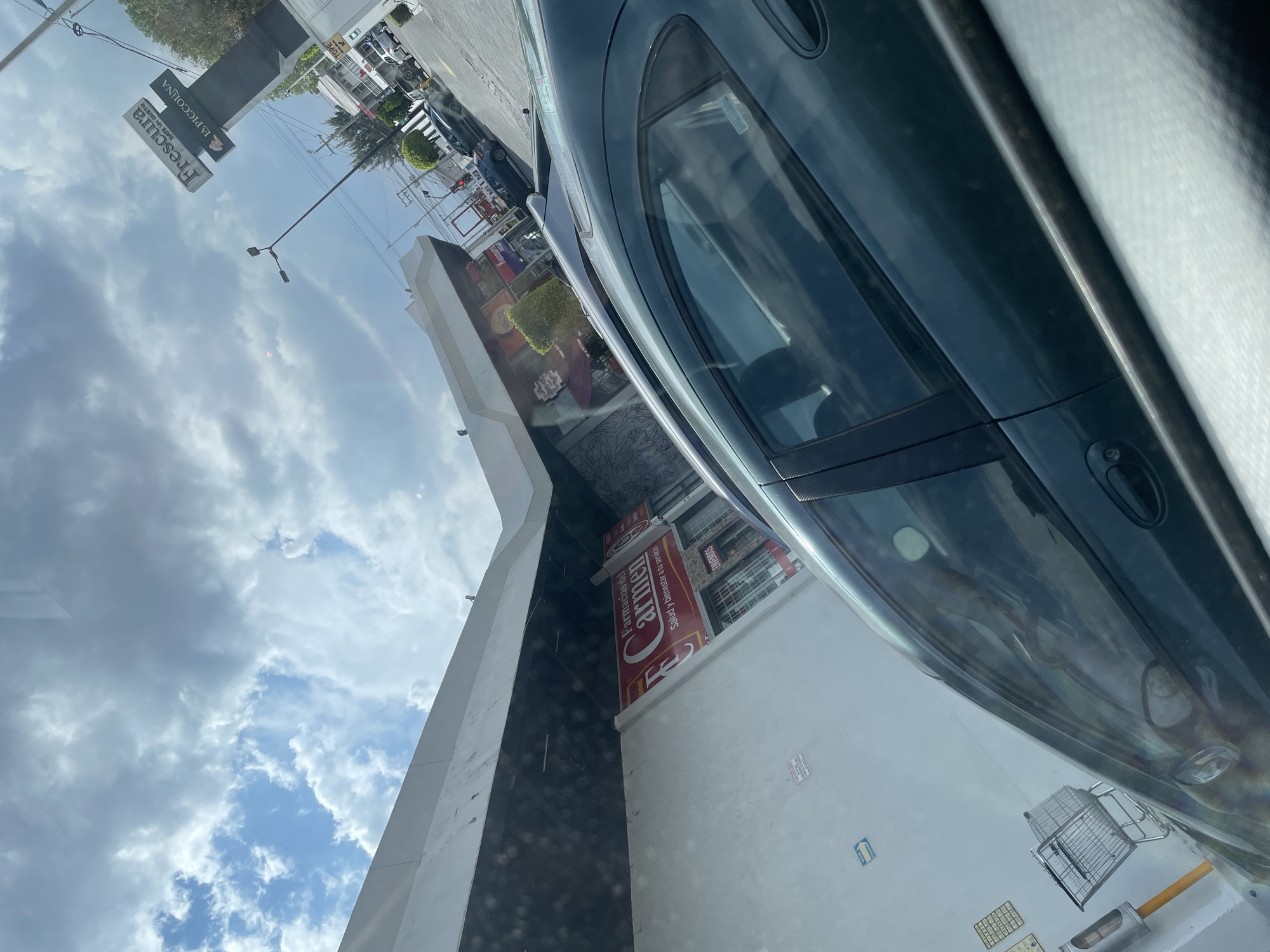 The image, taken from inside a car, shows a view of a street in what appears to be a commercial district. Through the car window, parts of a gray car are visible, with the reflection of the outside scene visible on the glass. Outside, the sky is cloudy, with patches of blue. A multi-story building is partly visible, housing establishments such as Frescura, La Piccolina, and Farmacias del Carmen. A sign reads "Salud y bienestar a tu servicio," which translates to "Health and wellness at your service". Other cars and traffic are also visible in the distance, indicating that this is an active commercial or retail area in the city of Puebla, Mexico. The angle and perspective of the photo suggest it was taken by someone sitting in the driver's seat of the car. There is also an empty shopping cart and some parking space marking tiles.