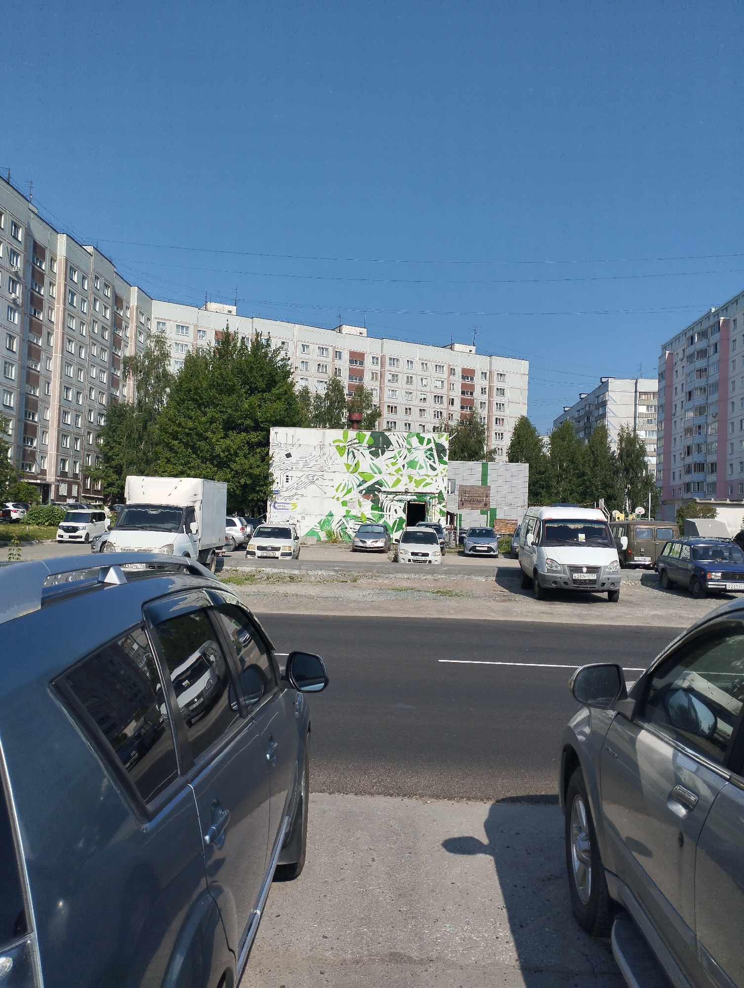 This is a full shot, eye-level, outdoor, daylight, medium-distance photograph taken in Novosibirsk, Russia. The image features a street with parked vehicles and apartment buildings in the background. On the left are multi-story apartment buildings with many windows, typical of Soviet-era architecture. In the middle ground, there is a parking lot with various vehicles including a white van, several passenger cars, and a larger van. The background is dominated by the sky and more apartment buildings. The overall setting suggests a residential area within a city.