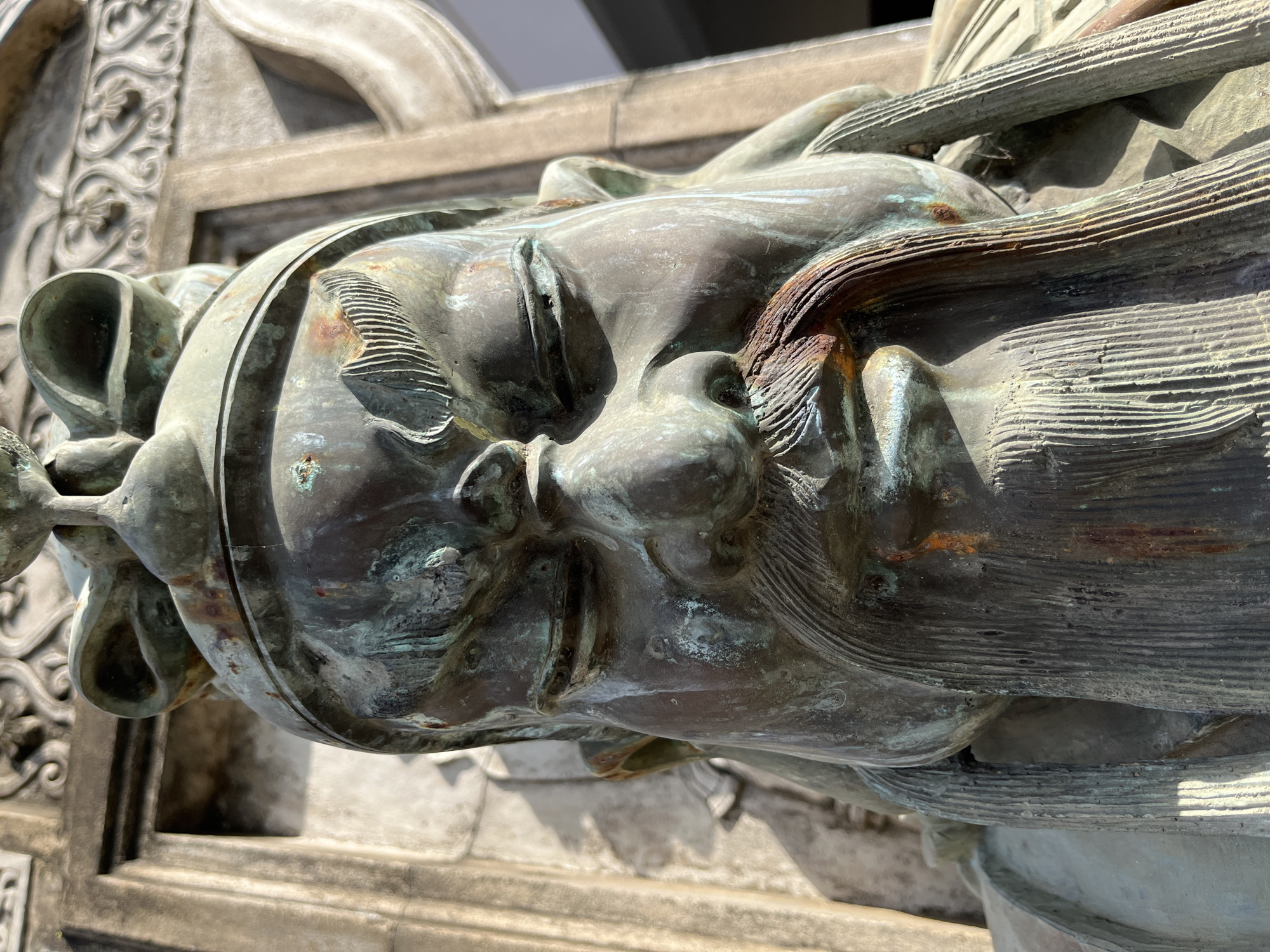 The image is a close-up, eye-level shot of a weathered bronze statue of a warrior, possibly Guan Yu, a Chinese deity. The statue, which is green with patina and hints of rust, depicts a stern-faced figure with closed eyes, a prominent mustache, and a long beard. The statue is situated in front of a stone structure with intricate carvings. The background is blurred, suggesting an outdoor setting. The statue is likely located at Gangaramaya Temple in Colombo, Sri Lanka, which is known for its eclectic collection of statues and artifacts.