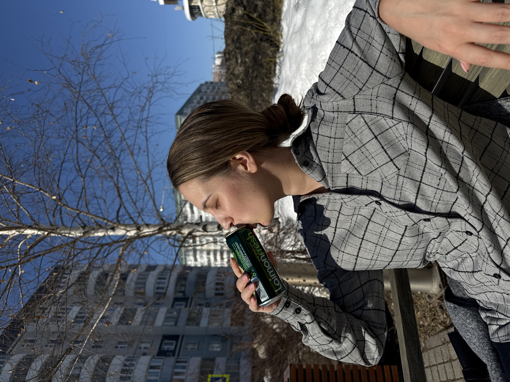 The image displays a young adult female seated on a dark wooden slatted bench under a clear blue sky in Kazan, Russia. She is shown in profile, eyes closed, drinking from a green aluminum can labeled "Laimon Fresh" with "Лимон Мята" (Lemon Mint) text visible. Her left hand holds the can, revealing light pink nail polish. She is wearing a grey long-sleeve button-up shirt with a black plaid pattern, and her brown hair is pulled back into a low bun.
The immediate environment includes patches of melting snow and dry ground, suggesting late winter or early spring. In the background, a bare-branched birch tree stands between a multi-story residential building with white balconies on the left and a large pile of dark earth or debris with snow patches on the right. Another building with a rounded upper section is visible in the distant right. A portion of a pedestrian crosswalk sign on a pole is visible in the left background.