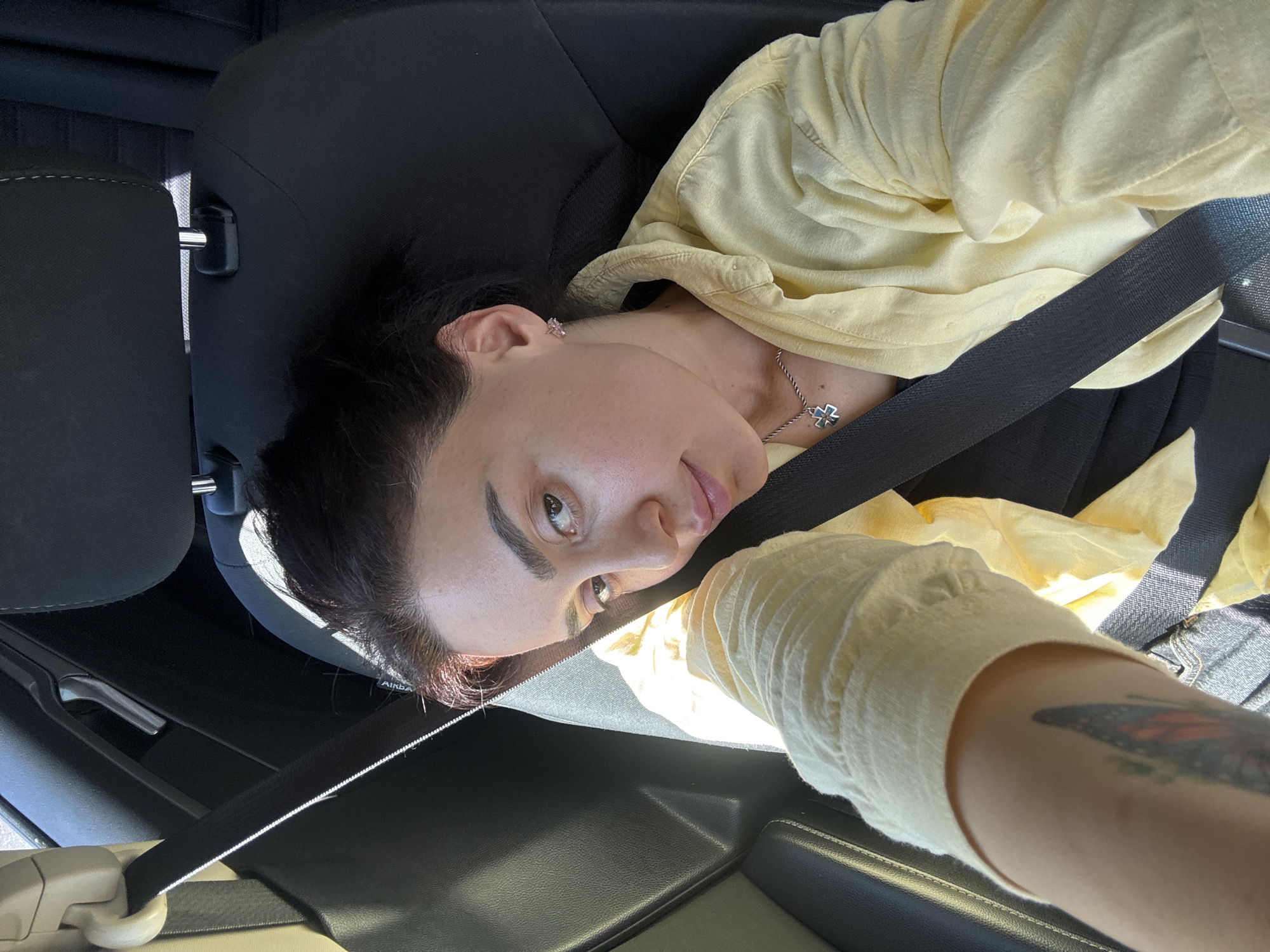 A woman is sitting in the driver's seat of a car, wearing a yellow shirt and a seatbelt. She has dark hair and a necklace. The car interior is visible, including the headrest of the seat. The location is unknown, as there are no visible landmarks to indicate it is Tbilisi, Georgia.
