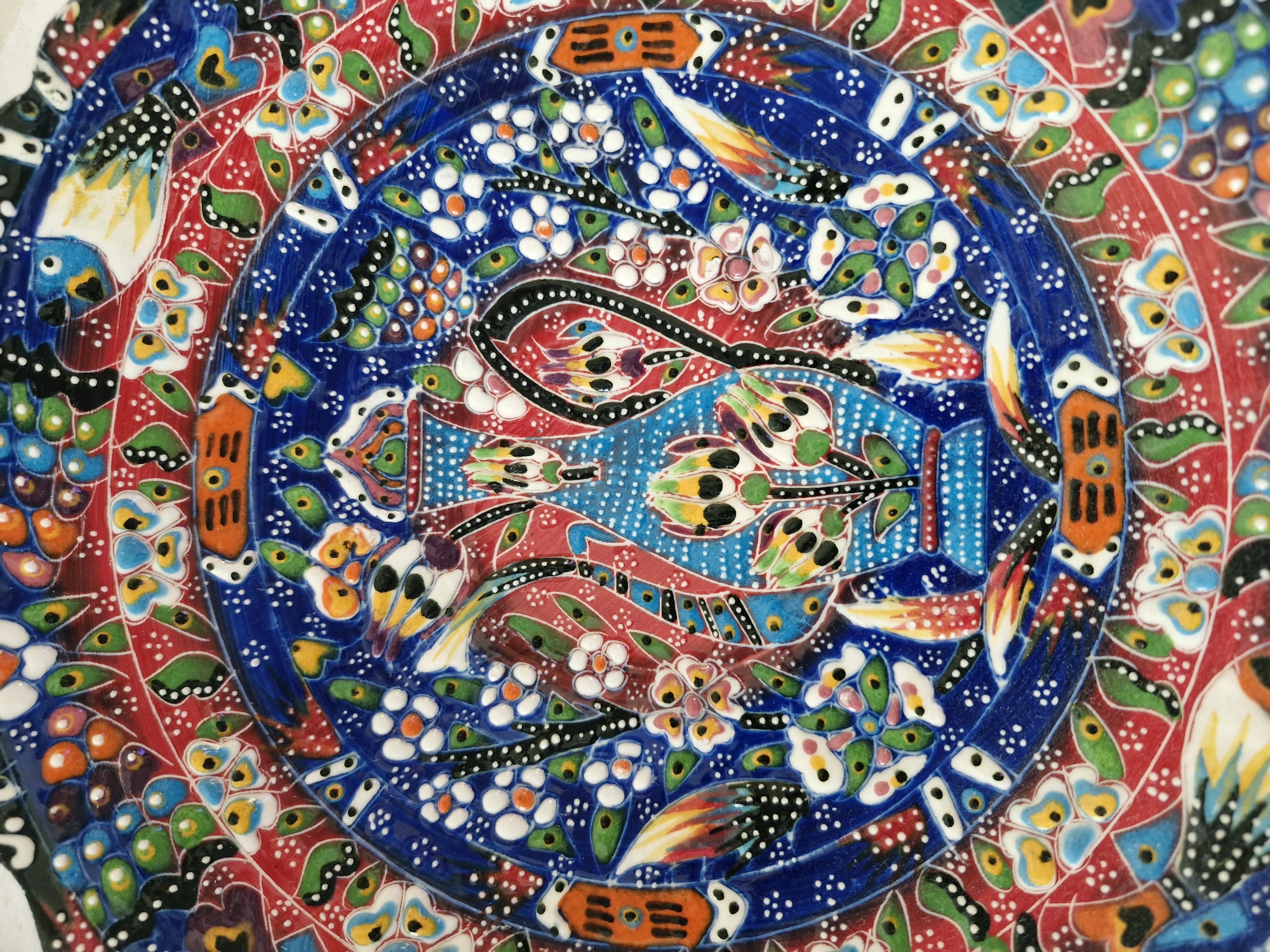 The image presents a close-up view of a circular, vibrantly colored ceramic plate or tile, showcasing an intricate and ornate design. The artwork is characterized by concentric rings of patterns and a rich palette of blues, reds, and whites, accented with green, yellow, orange, and black.
At the center, there is an elongated, stylized form, resembling either a decorative vase or a mythical creature, rendered in blue with red, black, and various multi-colored elements. This central figure is set against a deep blue background speckled with numerous small white dots. Surrounding this core motif is an inner ring of dark blue, densely decorated with white floral patterns and additional white dot accents.
Beyond this inner blue ring lies a broader, prominent red band. This red section is bordered by a wavy black line punctuated with white dots and is filled with a diverse array of abstract floral and leaf-like motifs in shades of green, yellow, orange, and light blue. Within this red band, stylized bird-like figures with yellow wings and blue bodies, along with other abstract fish or animal forms, are visible. The outermost visible sections continue with dark blue backgrounds adorned with white dots and further decorative patterns.
The ceramic surface appears glossy, and many of the painted details, particularly the white dots and certain colored areas, possess a slightly raised, textured quality, indicating an enamel or dimensional paint application. The overall aesthetic is highly decorative, reminiscent of traditional Turkish or Middle Eastern ceramic art. No visible text is present on the artwork.