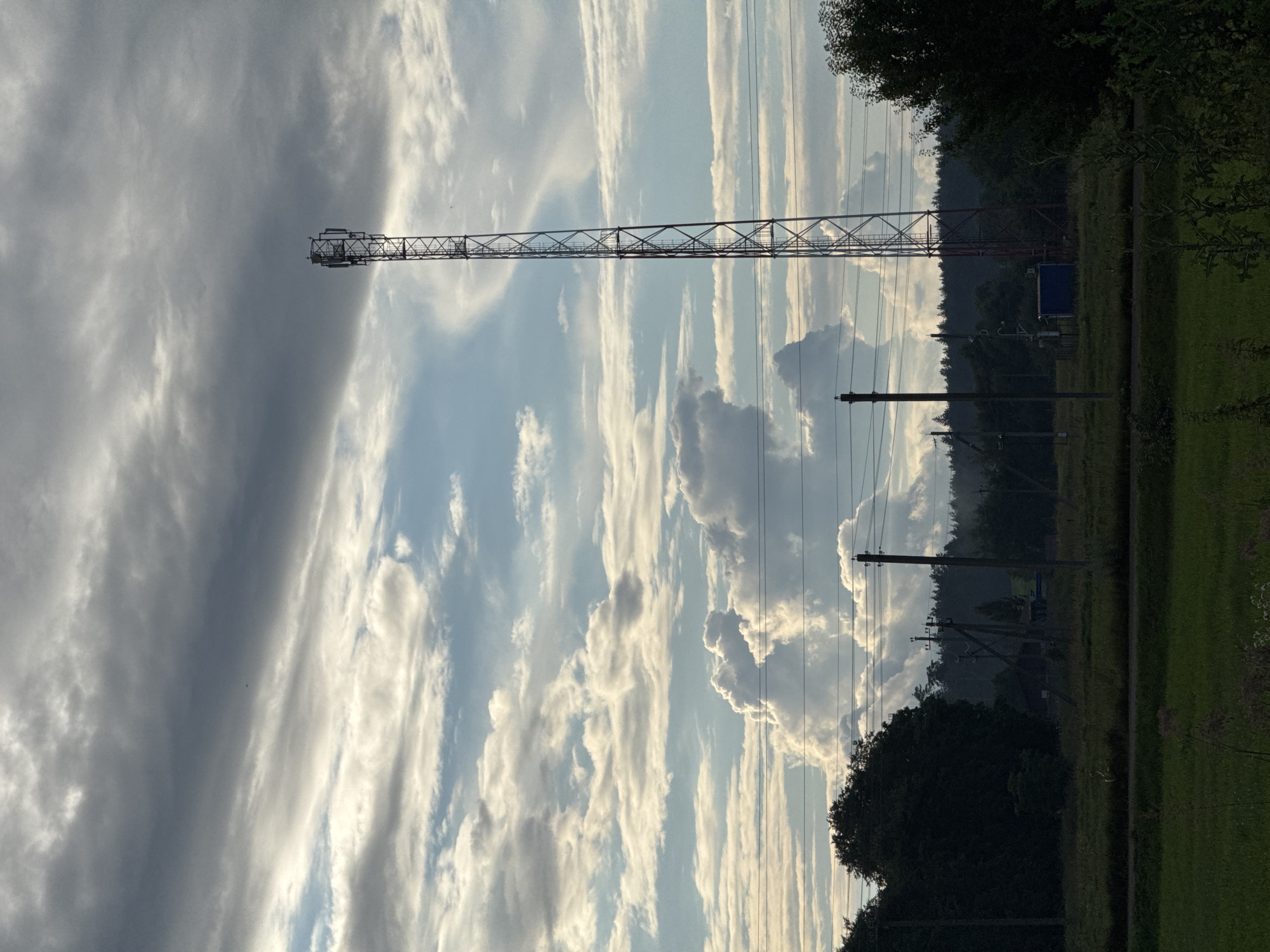 The image depicts a rural landscape under a dramatic sky. A tall, metallic telecommunications tower stands prominently in the mid-right portion of the frame, its structure silhouetted against the clouds. The sky is filled with a mix of dark, heavy clouds and patches of brighter, illuminated sky, creating a sense of dynamic weather. In the foreground, a dark, dense line of trees and vegetation runs across the bottom half of the image, with some power lines extending horizontally through the scene. Further back, more trees form a distant treeline, and several utility poles are visible. The ground appears to be grassy fields. The scene is likely located in the Нёманскі сельскі Савет of Belarus, a rural administrative division.