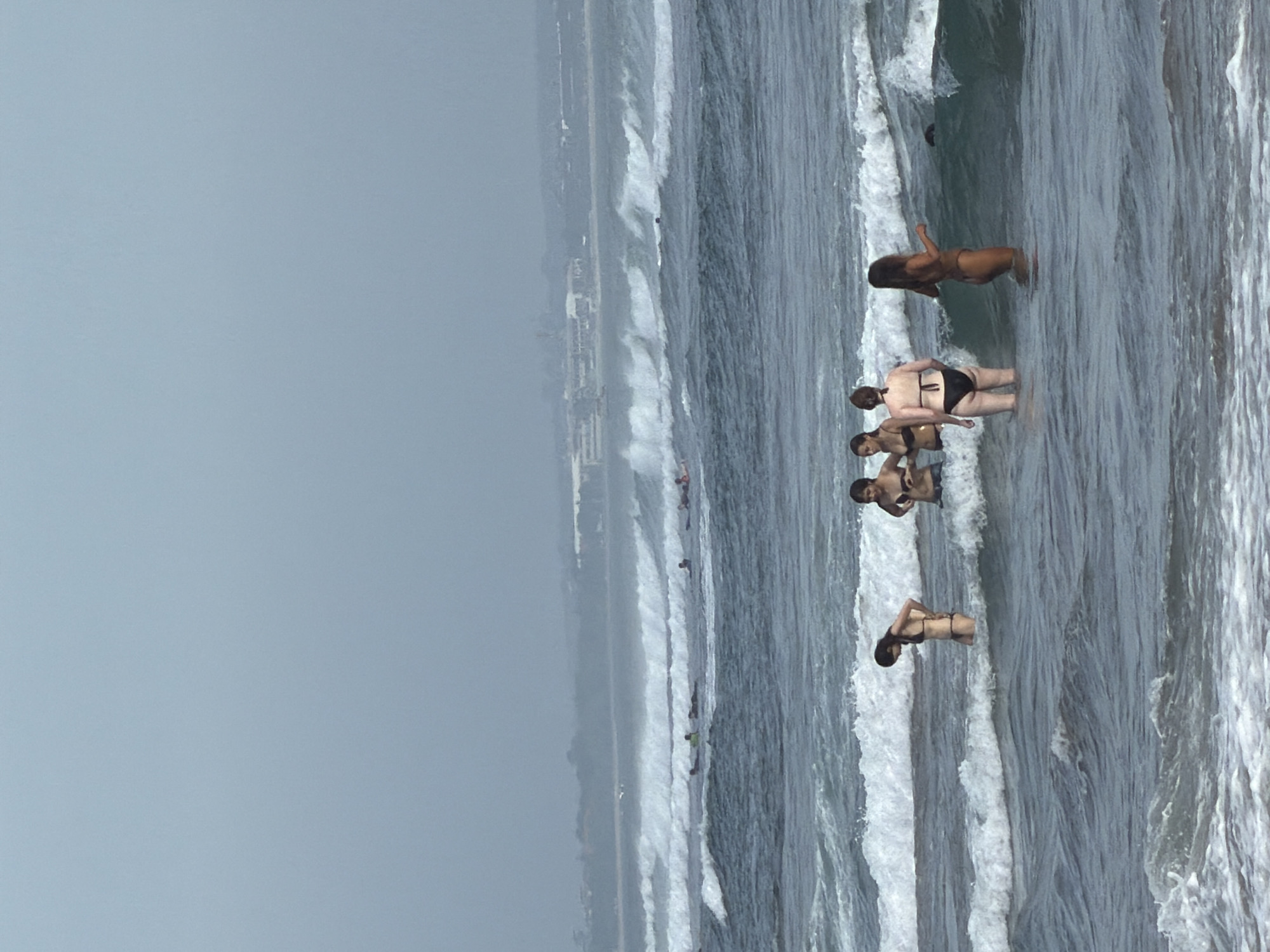 A group of people are in the ocean near the shore in North Kuta, Indonesia. Several individuals are standing or wading in the water, while others are further out on surfboards. The sky is overcast, and the ocean has moderate waves. In the background, a distant coastline with buildings is visible. The water is a murky blue-green color, and the waves are breaking white.