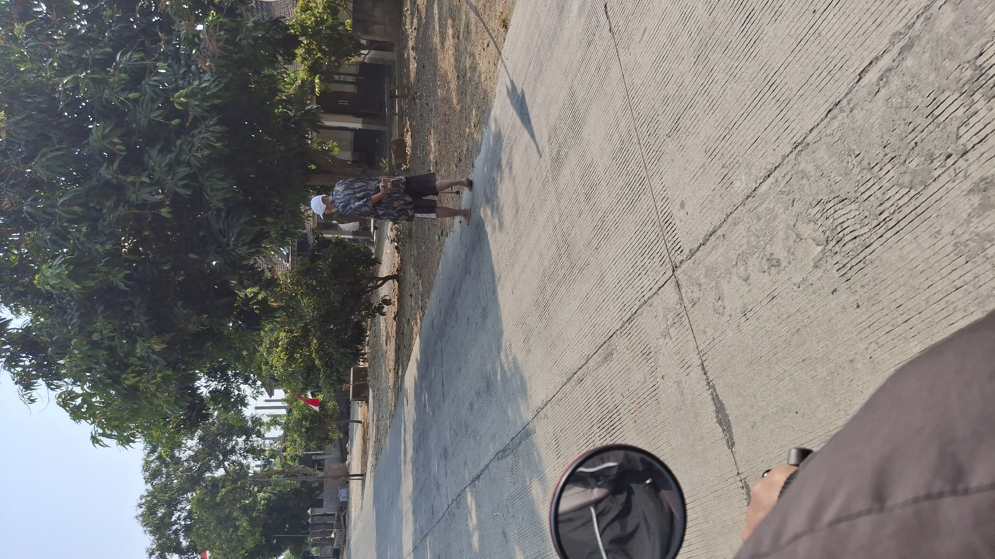 The image depicts a street scene in Pucakwangi, Indonesia. In the mid-ground, a person wearing a white cap, a patterned short-sleeved shirt, and patterned shorts is standing on the right side of a paved road, facing away from the camera. The person is barefoot and has their hands behind their back. The road is constructed of concrete with distinct parallel grooves. To the right of the person, there is a building with a light-colored facade and brown doors. A wooden bench is visible in front of the building. Various trees with green foliage are present in the background. In the left foreground, a portion of a motorcycle's handlebar and a side mirror are visible, suggesting the image was taken from a moving vehicle. Red and white flags are also visible in the background, attached to poles. The overall environment appears to be outdoors during the daytime with clear skies.