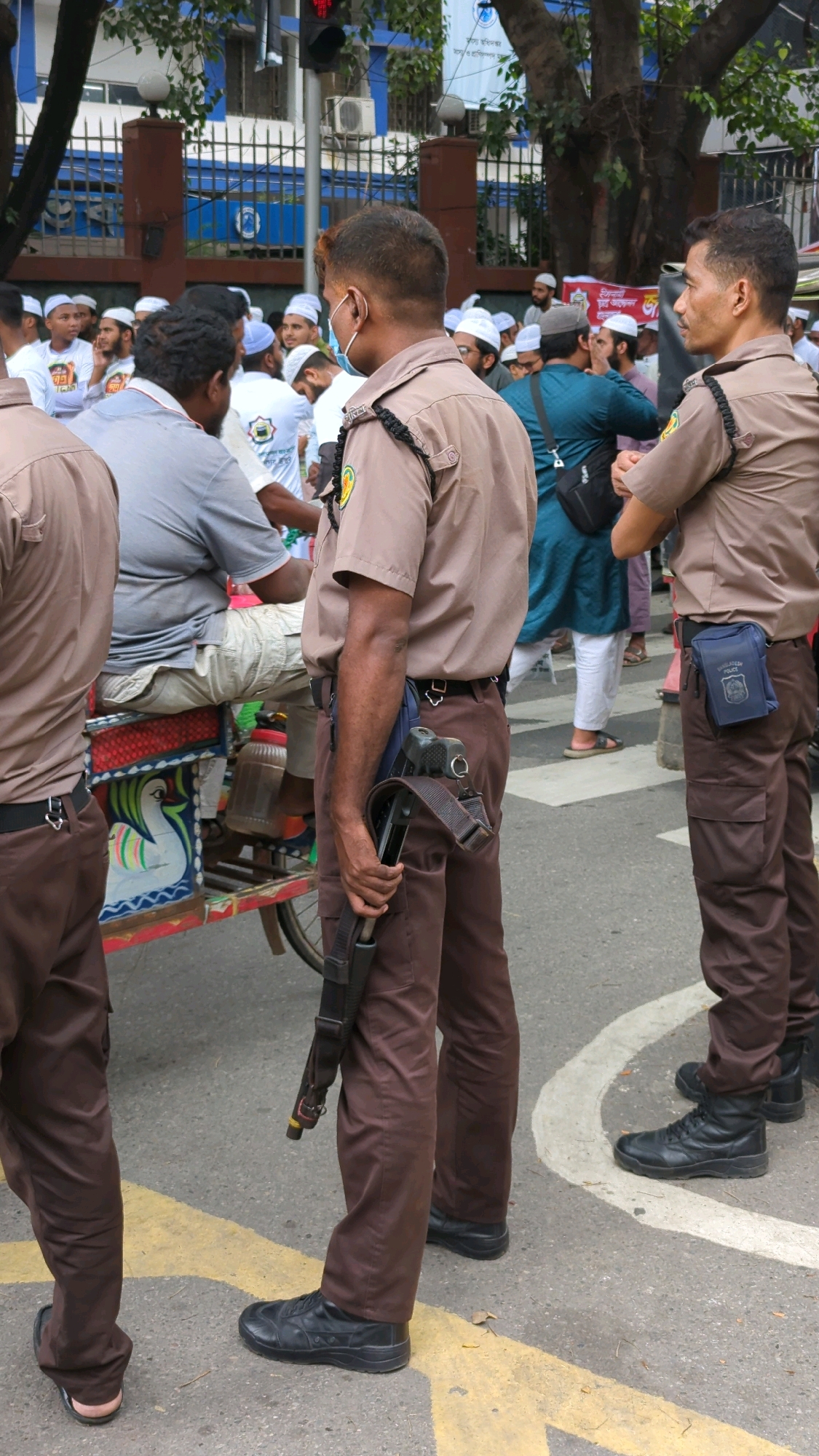 In Dhaka, Bangladesh, two men in uniform stand on a street. The man on the left, in the foreground, wears a light brown shirt and matching brown pants. He has a dark-colored firearm slung across his body, held in his right hand, with the barrel pointing downwards. He has a black belt with a silver buckle and black shoes. He is wearing a white face mask.
To his right, another man in a similar uniform stands facing away from the camera, also wearing a light brown shirt and brown pants. He has a blue pouch attached to his belt on his right hip. He is wearing black shoes.
Behind them, a crowd of people, many wearing white caps and shirts, are visible. One person is seated on a colorful cart. The background shows a building with blue and white signage and a black fence. Trees are present, and the ground is paved with asphalt and marked with yellow and white lines. The scene appears to be outdoors in a public area.