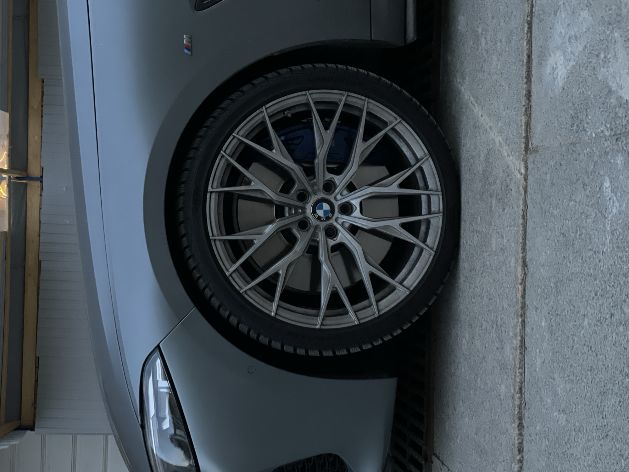 A close-up, angled shot depicts the front wheel of a gray BMW car. The wheel has a complex, multi-spoke design with a prominent BMW logo at the center. Visible behind the spokes are blue brake calipers. A small "M" badge is visible on the car's body near the wheel well. The car is parked next to a light gray concrete sidewalk. The setting is unidentified but the query asks it to be Saint Petersburg, Russia.