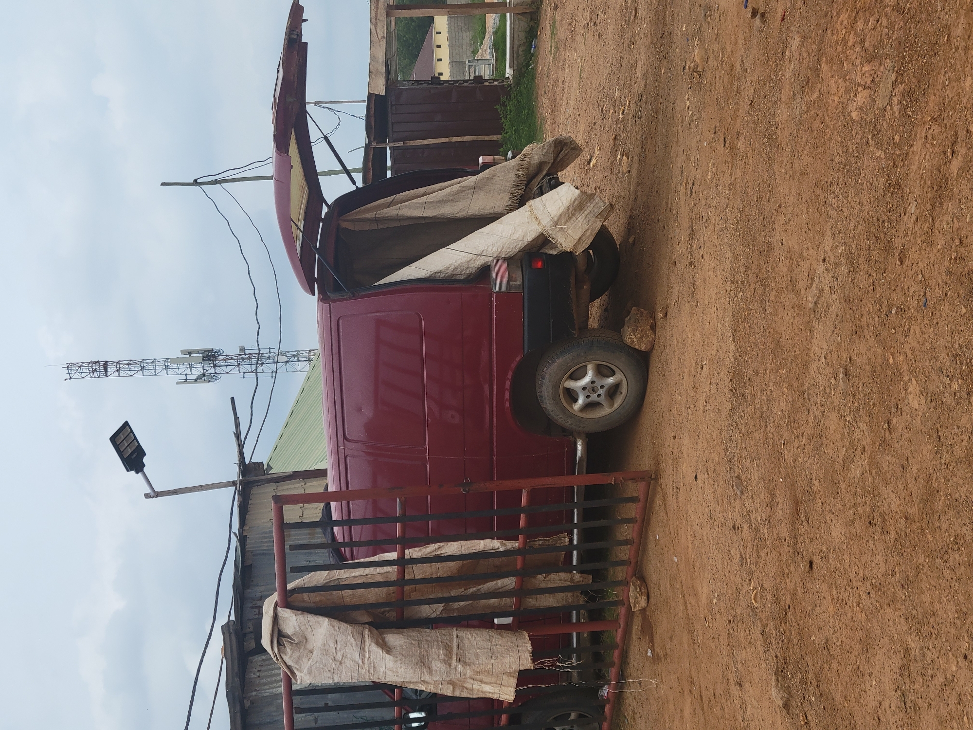 A burgundy van with its rear hatch open is parked on a dirt lot in Keffi, Nigeria. Large beige tarps are draped over the open hatch and the side of the van, obscuring its contents. A black and red metal gate, partially opened, stands in front of the van. In the background, a cellular tower rises above a cluster of corrugated metal structures and a partially constructed building with a green roof. The sky is overcast.