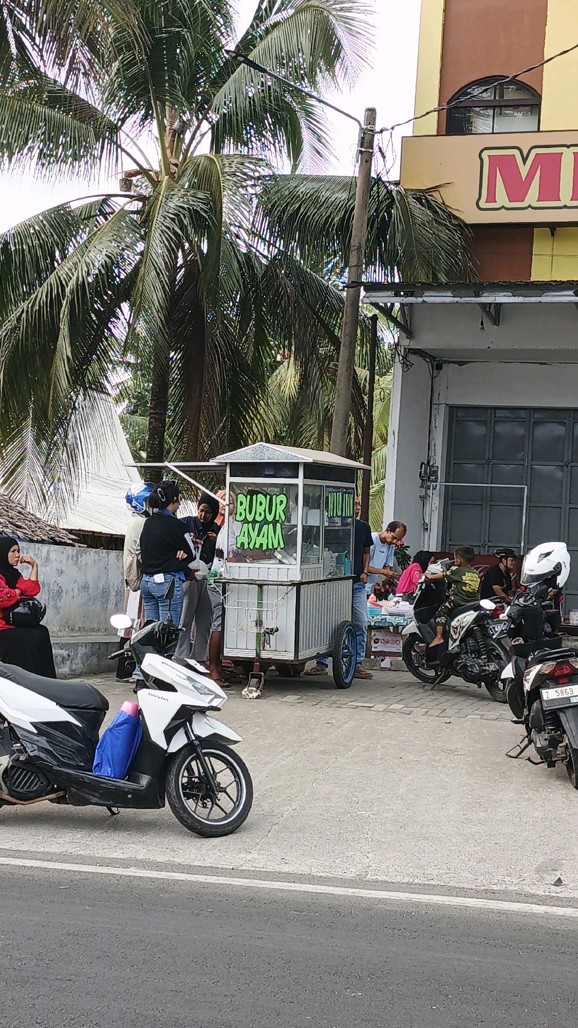 In Desa Wonoharjo, Indonesia, a food cart selling "BUBUR AYAM" (chicken porridge) is parked on a sidewalk adjacent to a building. Several people are gathered around the cart, with some appearing to be customers and others likely vendors. Two motorcycles are parked on the sidewalk in the foreground, one white and one black. The building behind the food cart has a sign that partially reads "MI". Palm trees are visible in the background. The scene appears to be a typical street scene in a commercial area.