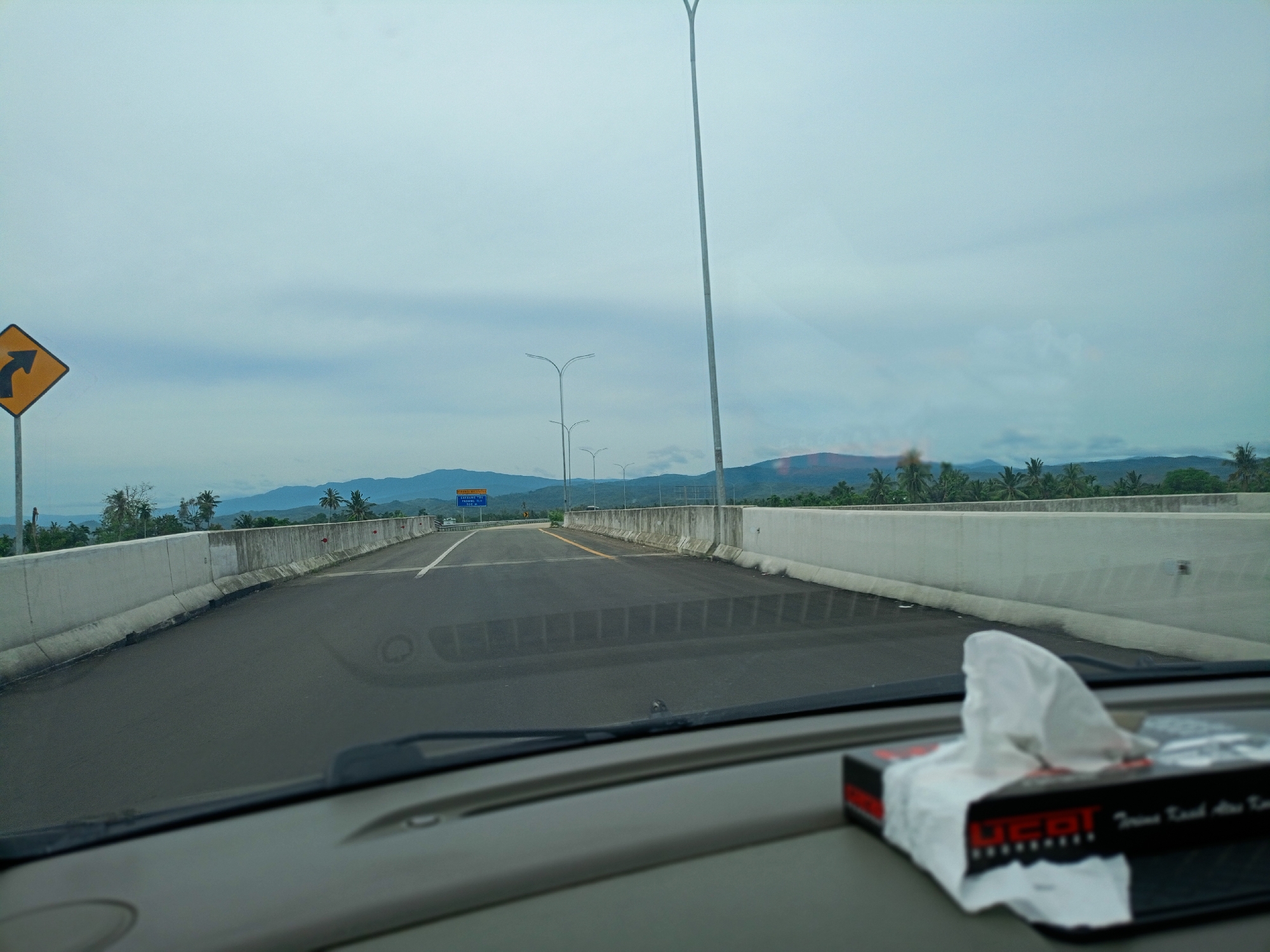The image shows a view from inside a car driving on a bridge or elevated road in Mukee Gogo, Indonesia. The road is paved and has concrete barriers on either side. There is a sign with a yellow background and a curved arrow pointing left on the left side of the road. A row of light poles lines the road ahead. In the distance, there are mountains under a cloudy sky. The car's dashboard is visible in the foreground, with a box of tissues on it. The windshield and wipers are also partially visible.