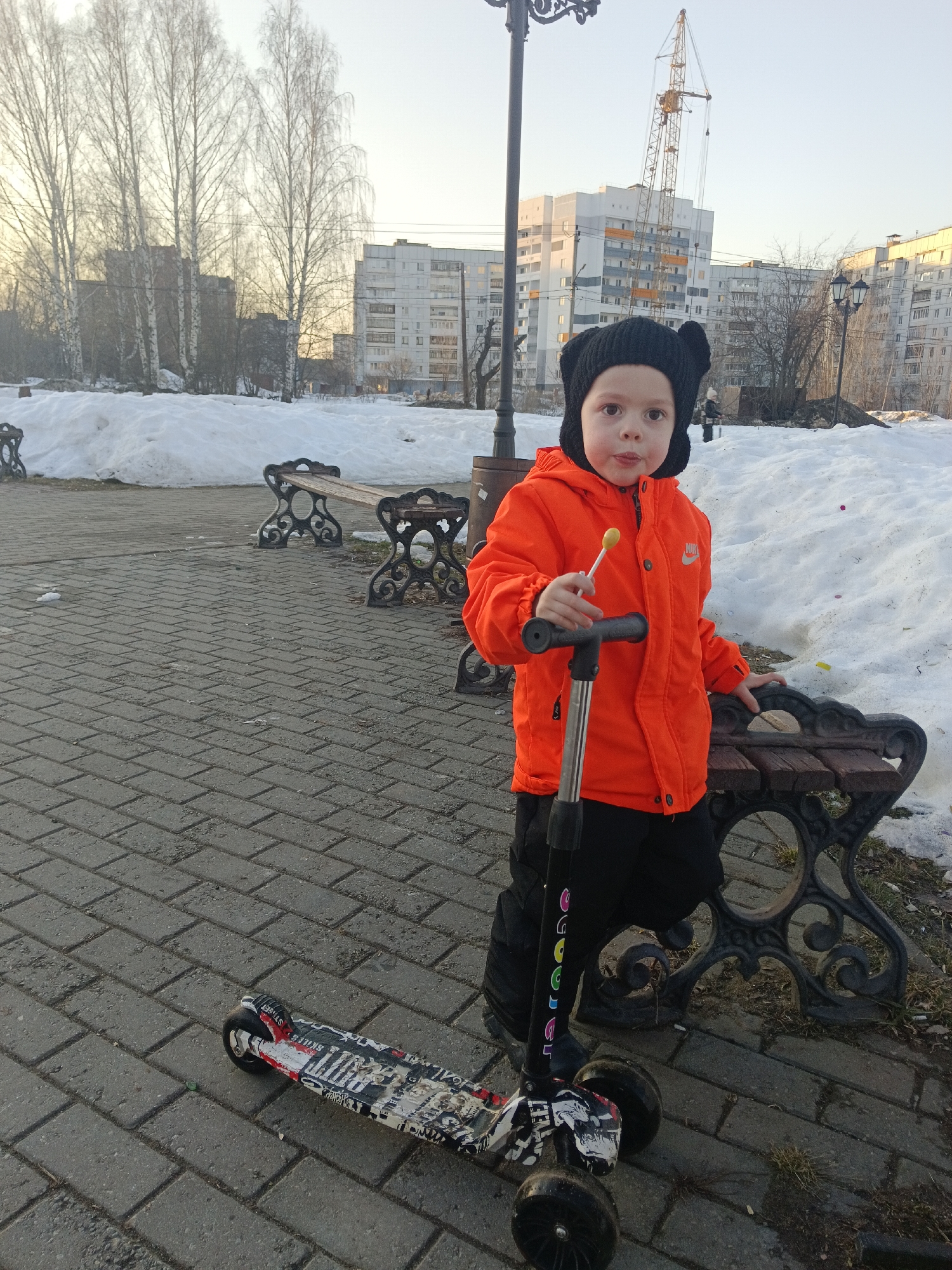 A young child stands centrally on a grey brick-paved path in a park setting within the city of Chaikovsky, Russia. The child, wearing a black ear-flapped hat, an orange Nike jacket, and black pants, holds a yellow lollipop stick to their mouth with their right hand and grips a three-wheeled scooter's handlebar with their left. The scooter's deck is adorned with graffiti-style graphics. The child's right hand rests on the ornate cast-iron armrest of a dark wooden park bench situated to their immediate right.
Behind the child and benches, large piles of melting snow line the edges of the paved area, interspersed with patches of dry grass and dirt. Several bare deciduous trees, possibly birches, stand in the midground, with branches reaching towards a bright, clear sky. In the background, multiple multi-story residential buildings are visible. One prominent white and grey building, appearing to be under construction or recently completed, is flanked by a large yellow tower crane. Other similar apartment blocks are scattered across the urban horizon. An ornate black lamppost is partially visible to the child's left, and another is seen further back on the right. The scene suggests late winter or early spring conditions with low sunlight.