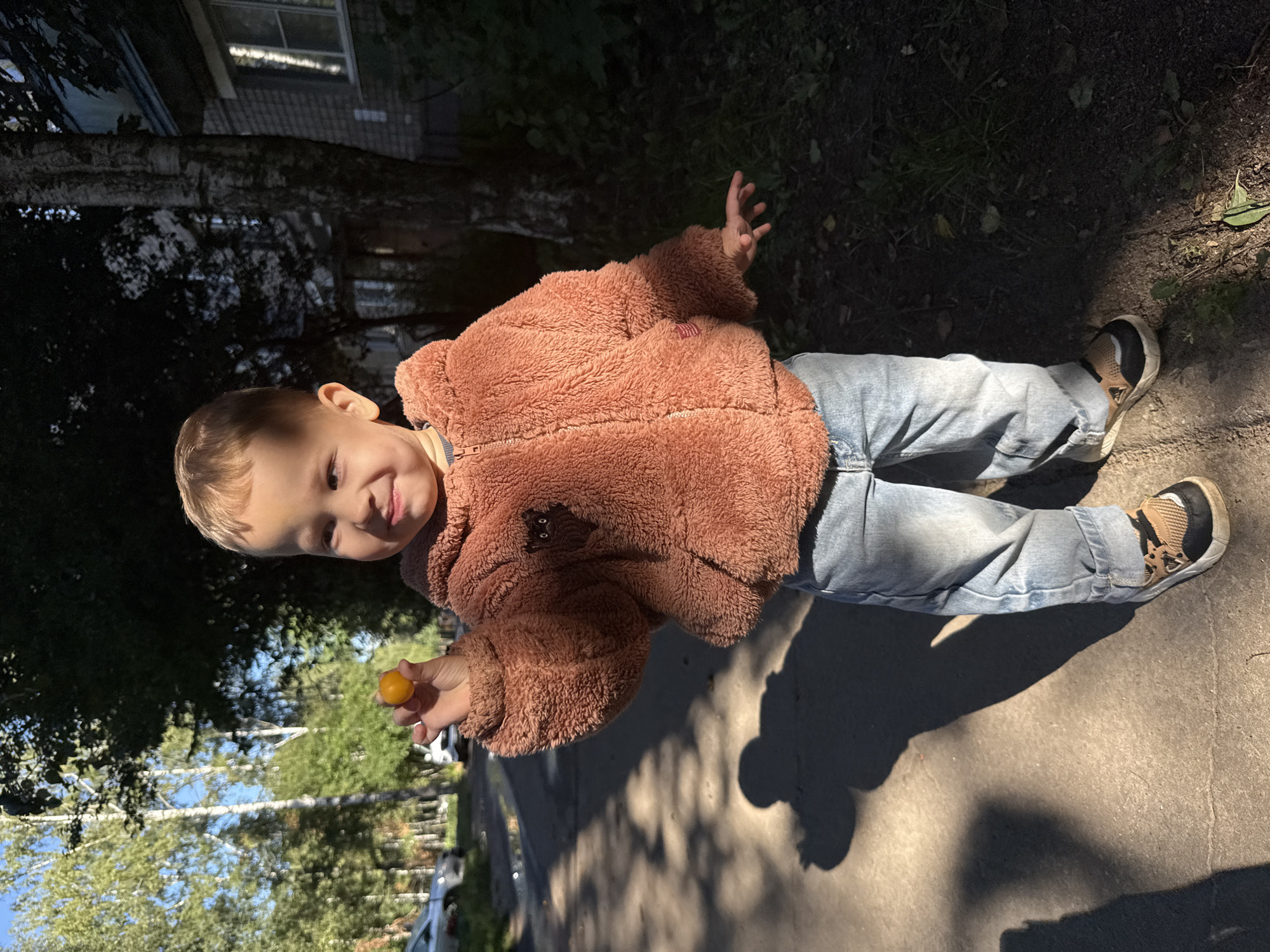 A young boy stands on a paved path in front of a building, holding a small orange object, possibly a cherry tomato, in his right hand. He is wearing a fluffy rust-colored jacket and light blue jeans. The scene is outdoors, with trees and shadows visible. The background includes a building with windows and a tree with rough bark. The lighting suggests it is daytime, with dappled sunlight. The context of Rusanovo, Russia is not visually apparent from the image.