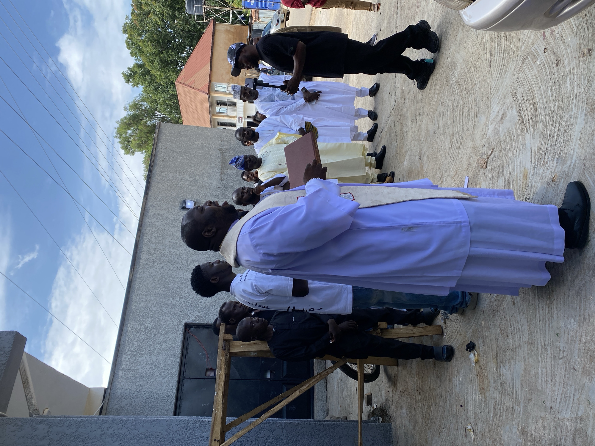 The image depicts an outdoor scene in Gura Top, Nigeria, likely near a residential or commercial building. A group of men, dressed in a mix of traditional and casual attire, are gathered on a concrete surface. In the foreground, a man in a light purple robe and stole, possibly a clergyman, is speaking and holding a book. Another man in a black t-shirt and cap is standing opposite him, holding a smartphone on a gimbal, appearing to record. The background shows a light gray building with a metal door, a wooden scaffolding structure, and a red-roofed building in the distance. Trees and a cloudy sky are visible above. The ground is littered with small debris. Several other men are observing the interaction, some in white traditional outfits. A motorcycle is partially visible behind the scaffolding. The overall atmosphere suggests a formal gathering or event.