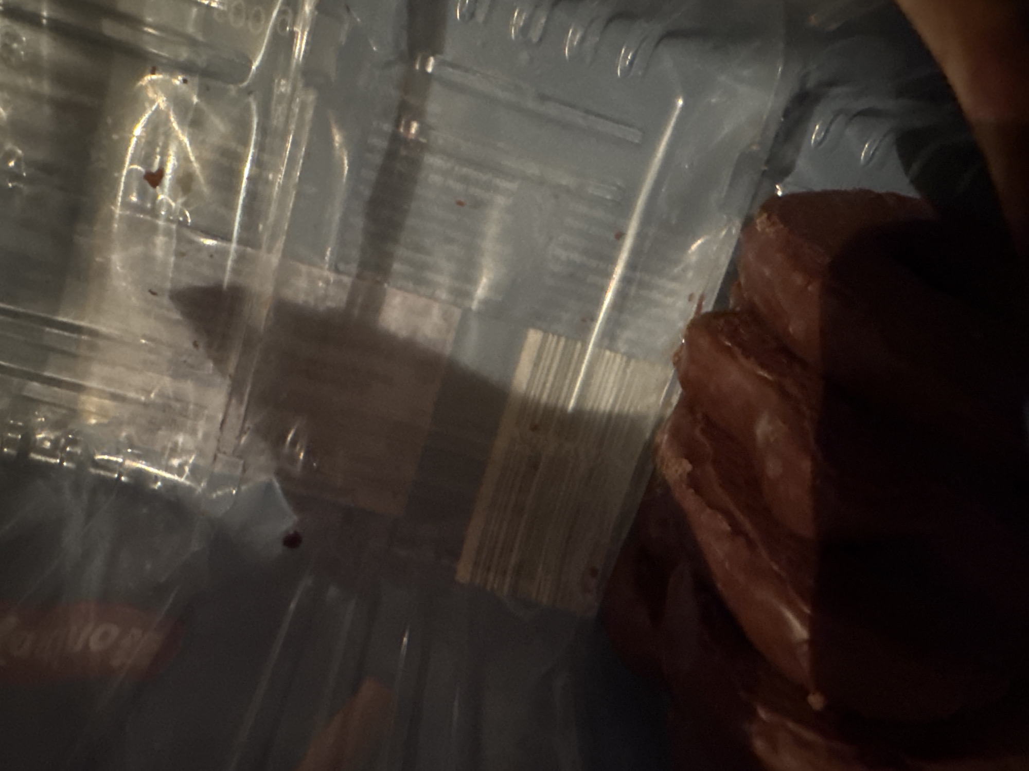 The image shows a close-up view of a dark red, processed meat product, possibly sausages or cured meats, packaged in clear plastic. The product is positioned in the foreground, with the plastic packaging and some internal structure of the packaging visible in the background. There are some dark red spots or smudges on the packaging. The lighting is dim, casting shadows and highlighting the texture of the meat. The scene appears to be indoors, likely within a kitchen or retail environment. There is no indication of the location being in Ruzhitsa, Bulgaria.