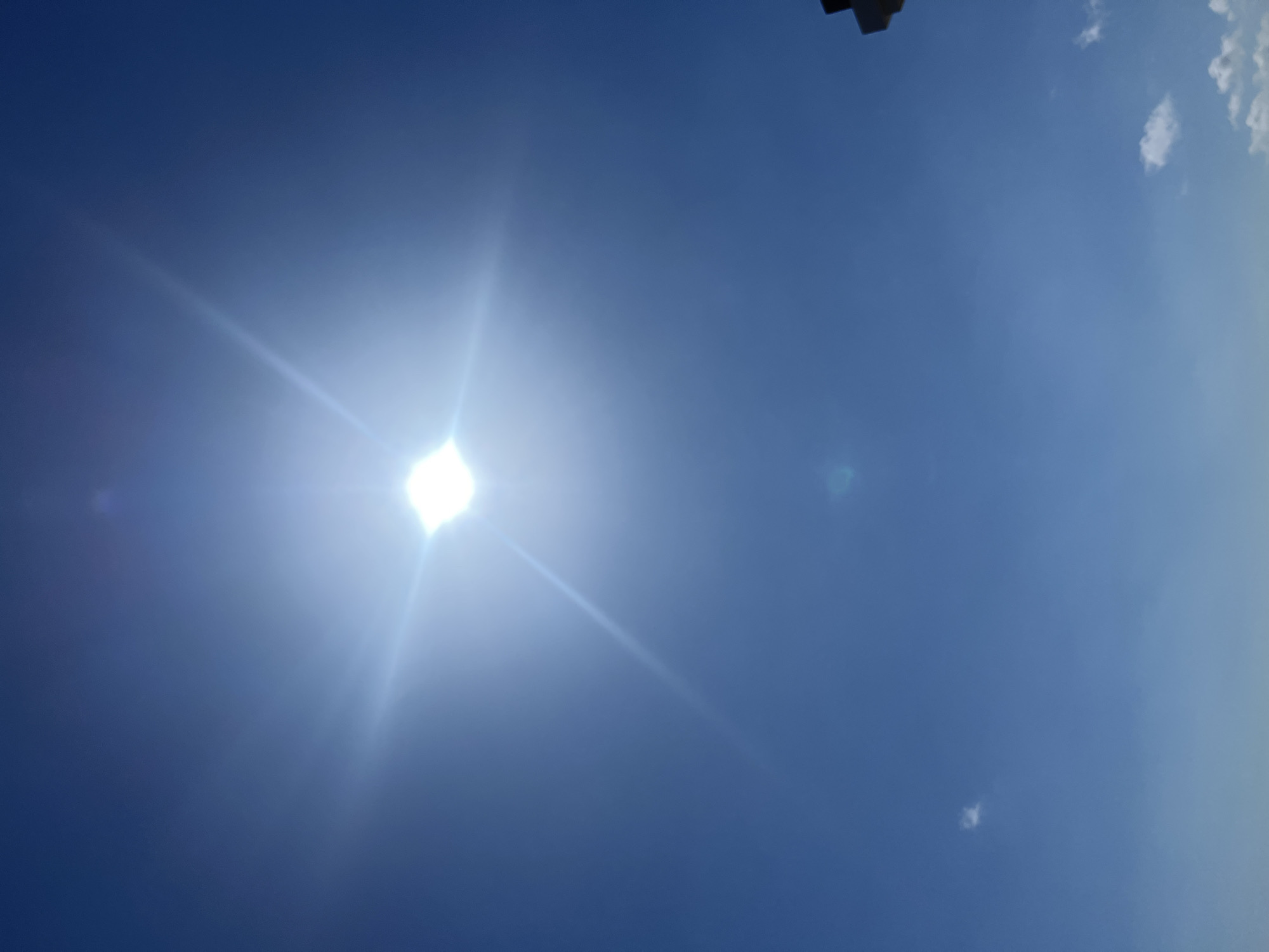 The image shows a bright sun in a clear blue sky, with visible lens flare. There are a few small white clouds scattered in the sky. In the bottom right corner, the top of a dark, angular object, possibly a building or antenna, is visible. The overall scene suggests a sunny day, likely outdoors. There is no indication of Kuta, Indonesia.