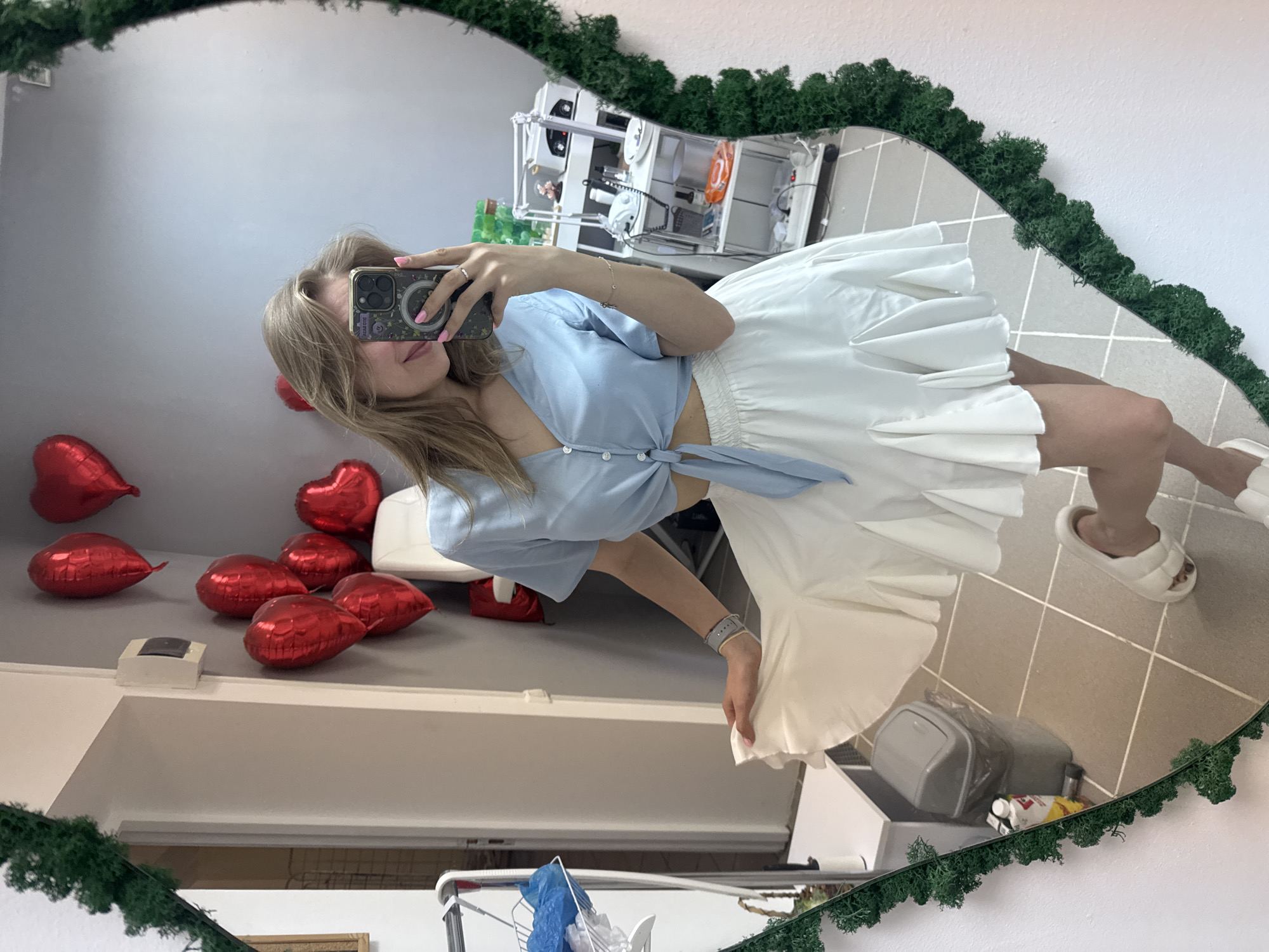 A woman is taking a selfie in a mirror with a green moss frame. The mirror reflects a bathroom setting, including red heart-shaped balloons, a white countertop, and various bathroom fixtures. The woman is wearing a light blue top tied at the waist and a white skirt. She is holding a phone up to her face. The reflection shows beige floor tiles and a trash can. The mirror's reflection contains the interior of a home and includes objects like a laundry rack, and various containers under the sink. There is no evidence or indication of location in Izhevsk, Russia.