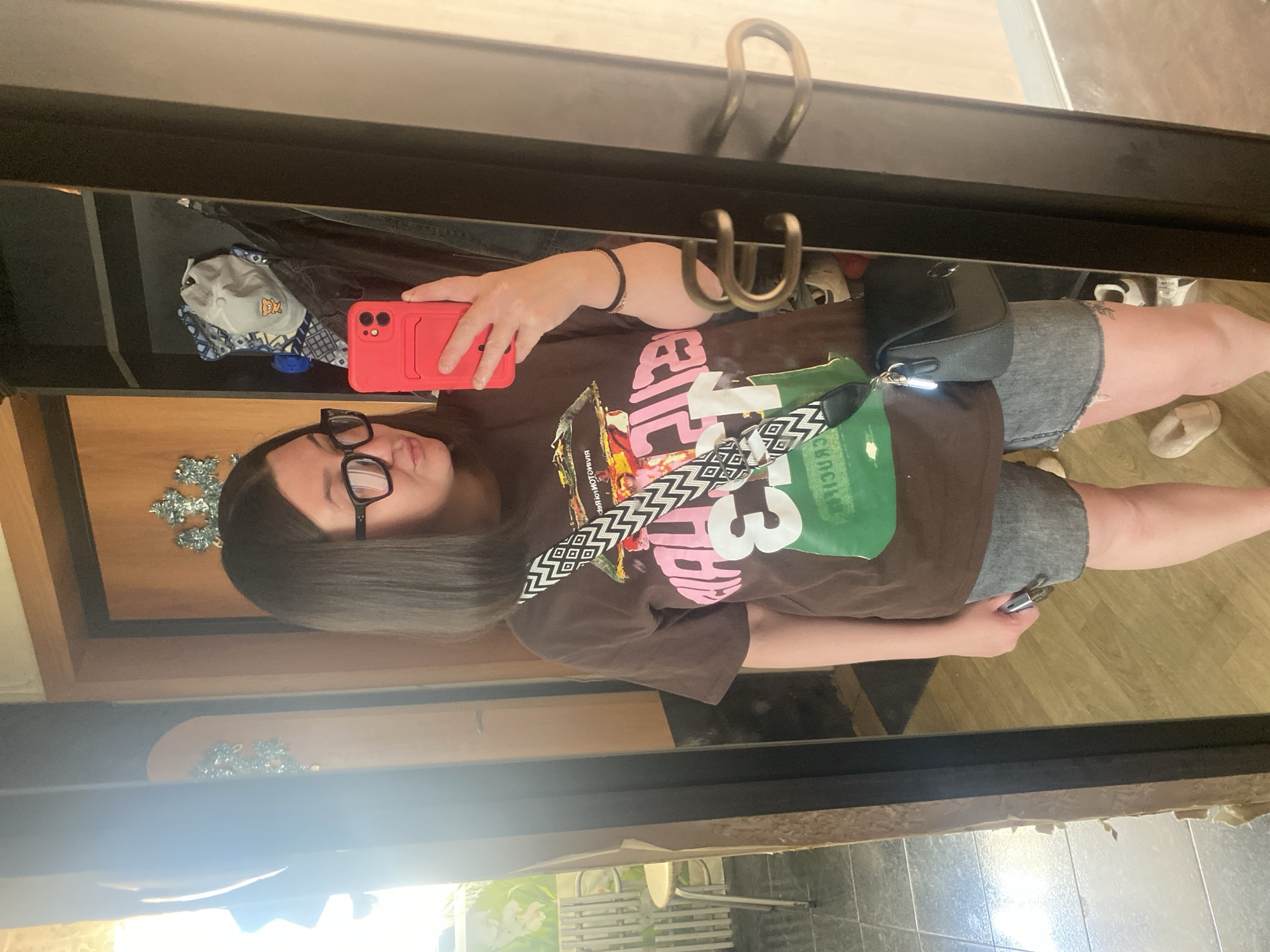 A person wearing glasses and a brown t-shirt with graphic designs is taking a selfie in a mirror. The person has a black shoulder bag with a black and white strap. The mirror reflects a glimpse of the surrounding room. The location is Chaikovsky, Russia, based on the Russian text visible in the graphic design of the t-shirt, specifically the word "Чайковский". The floor appears to be a wood-look tile.