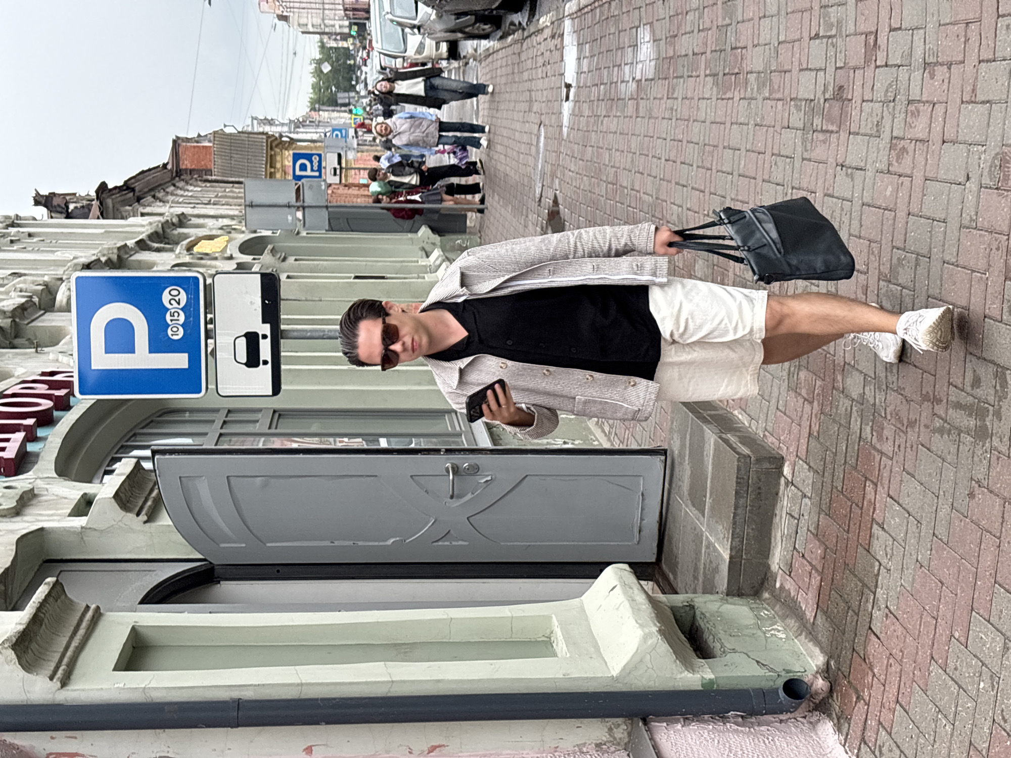 A man with dark hair and sunglasses walks along a brick sidewalk in Nizhny Novgorod, Russia, while looking at his smartphone. He is wearing a light-colored, pinstriped jacket over a black button-up shirt and white shorts. He carries a black tote bag in his right hand. The sidewalk is wet, suggesting recent rain. Behind him, other pedestrians walk further down the street. Buildings line the left side of the sidewalk, one with a light green facade and an open gray door. A blue parking sign with white lettering is visible on a pole in the background. Cars are parked along the right side of the street.