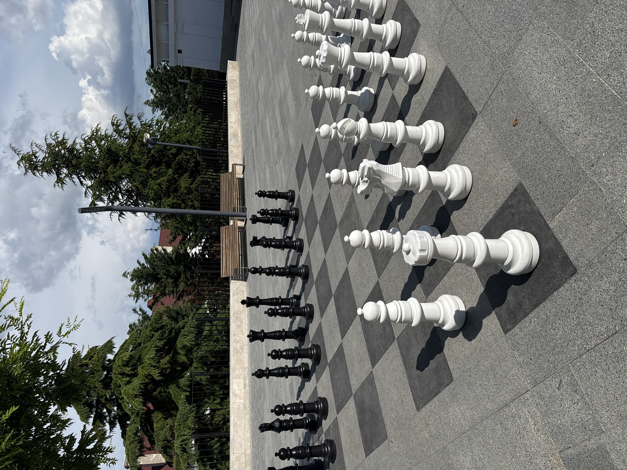 An outdoor giant chess set is set up on a checkered pavement. The chess pieces are white and black, and they are arranged as if in the middle of a game. The white pieces are on the right side of the image, and the black pieces are on the left side. In the background, there are trees, a fence, and buildings. The sky is cloudy.