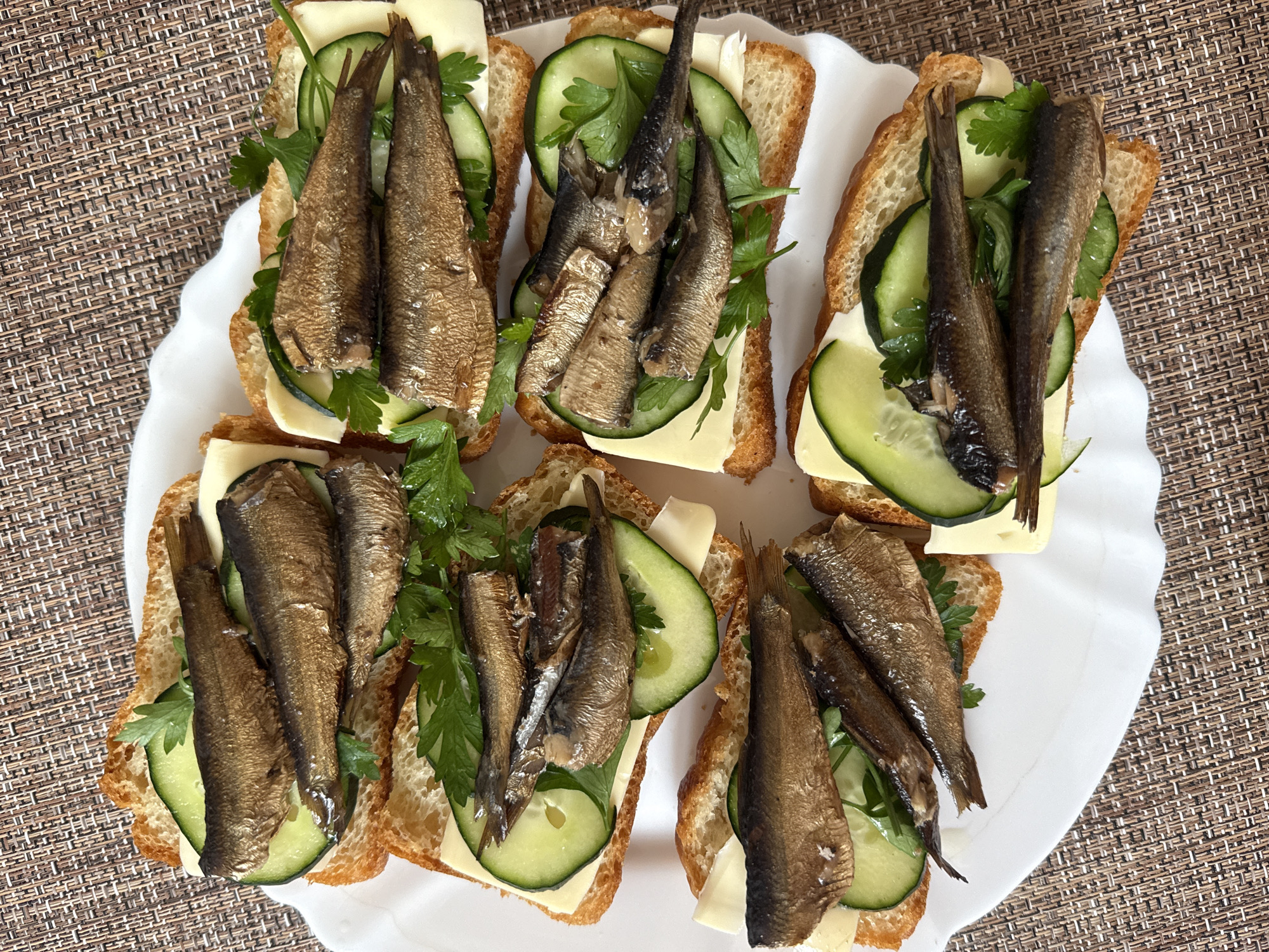 Six open-faced sandwiches are arranged on a white plate. Each sandwich consists of a slice of toasted bread topped with cheese, slices of cucumber, parsley, and several canned sardines. The plate is set on a woven placemat. The scene is a close-up overhead shot of the food.