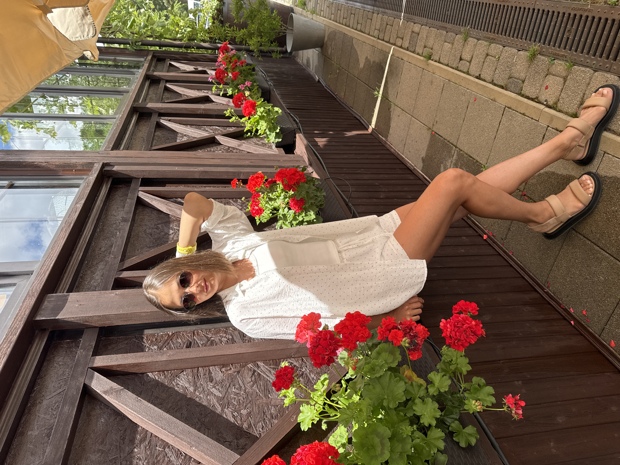 A young woman with blonde hair, wearing sunglasses, a white polka-dot shirt, white shorts, and tan sandals, leans against a wooden structure with a planter box of red flowers. She has her right arm raised behind her head. The scene appears to be an outdoor seating area, possibly at a cafe, in Abrau-Dyurso, Russia, as indicated by the presence of grapevines visible in the background and the architectural style suggestive of a winery or resort. The wooden structure is stained dark brown and features a lattice design. The planter box contains lush green foliage and bright red flowers, with some petals scattered on the cobblestone walkway. A beige awning is visible overhead.