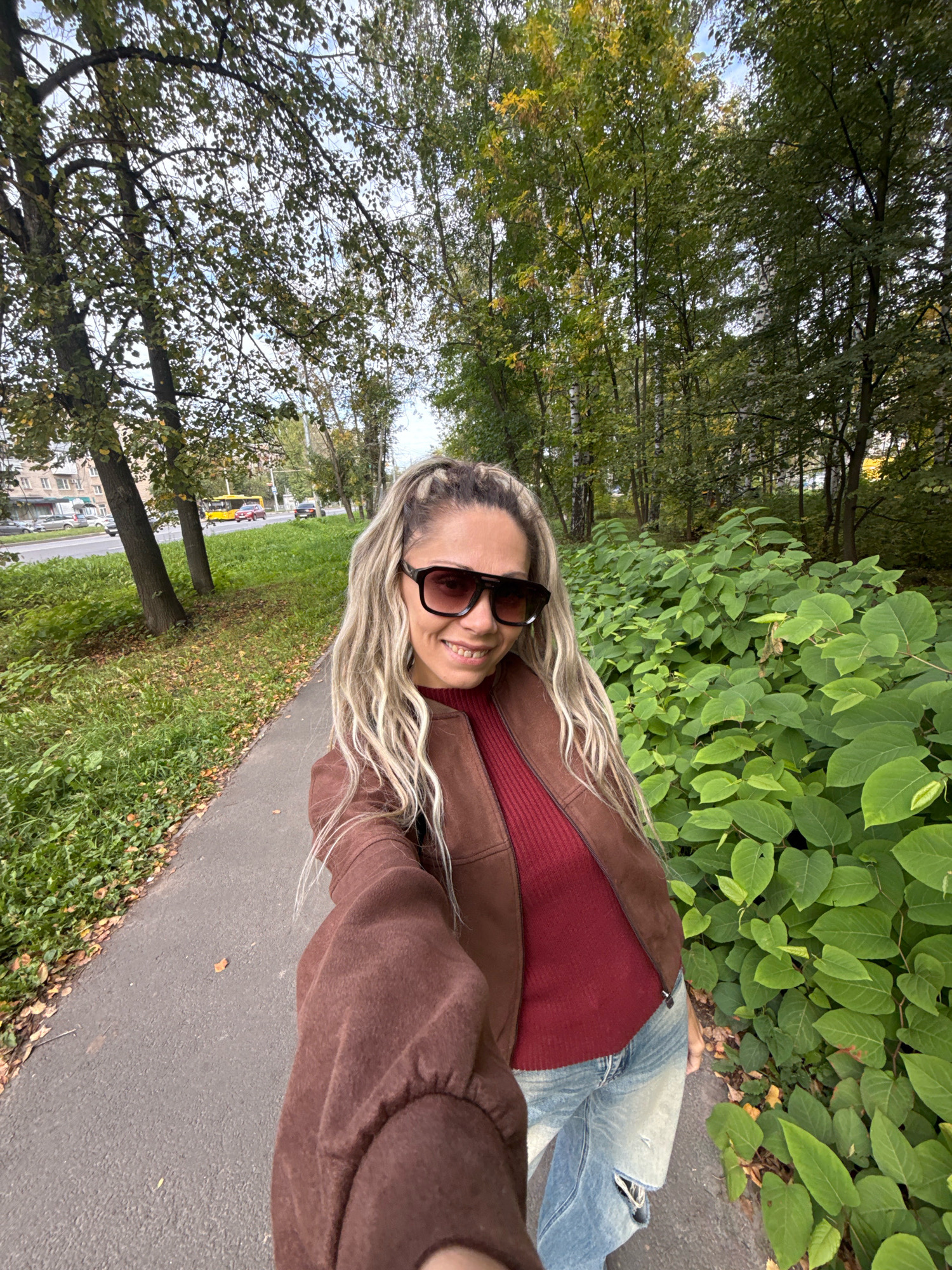 A woman with blonde hair pulled back in a ponytail, wearing sunglasses, a brown jacket, a maroon shirt, and ripped jeans, smiles at the camera as she takes a selfie on a paved path lined with green plants and trees. To the left of the path, there is a grassy area with fallen leaves and a road with cars and a yellow bus. The background shows more trees and buildings. The scene appears to be in an urban park or along a tree-lined street in Rusanovo, Russia.