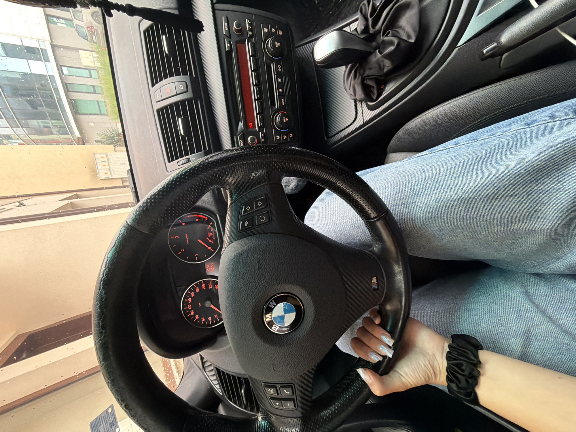 The image provides a first-person perspective from the driver's seat of a car in Gabrovo, Bulgaria, looking forward through the windshield. The driver, wearing light blue jeans, has their right hand gripping a black, carbon-fiber textured steering wheel, which prominently features a BMW logo in the center and an "M" logo on the right spoke. The driver's hand has long, light-colored fingernails, and a black scrunchie is worn on the wrist.
Visible on the dashboard are two analog gauges with red backlighting: a speedometer showing speeds up to "240" and a tachometer indicating RPMs up to "5" (x 1000). A digital display on the dashboard shows "12:59" for the time and "23.5°C" for the temperature. The center console includes a radio and climate control unit with buttons labeled "MENU," "AUDIO," "AUTO," and "REST," alongside air vents and control knobs. A gear shifter and handbrake are also visible to the right of the driver. The car's interior features carbon fiber-like trim accents on the console.
Through the rain-speckled windshield, the exterior scene shows modern buildings with large glass windows, indicating an urban or suburban setting. The presence of raindrops suggests recent or ongoing rain. The time display of "12:59" points to midday. The image captures a paused moment within the vehicle, possibly during a stop or parking.