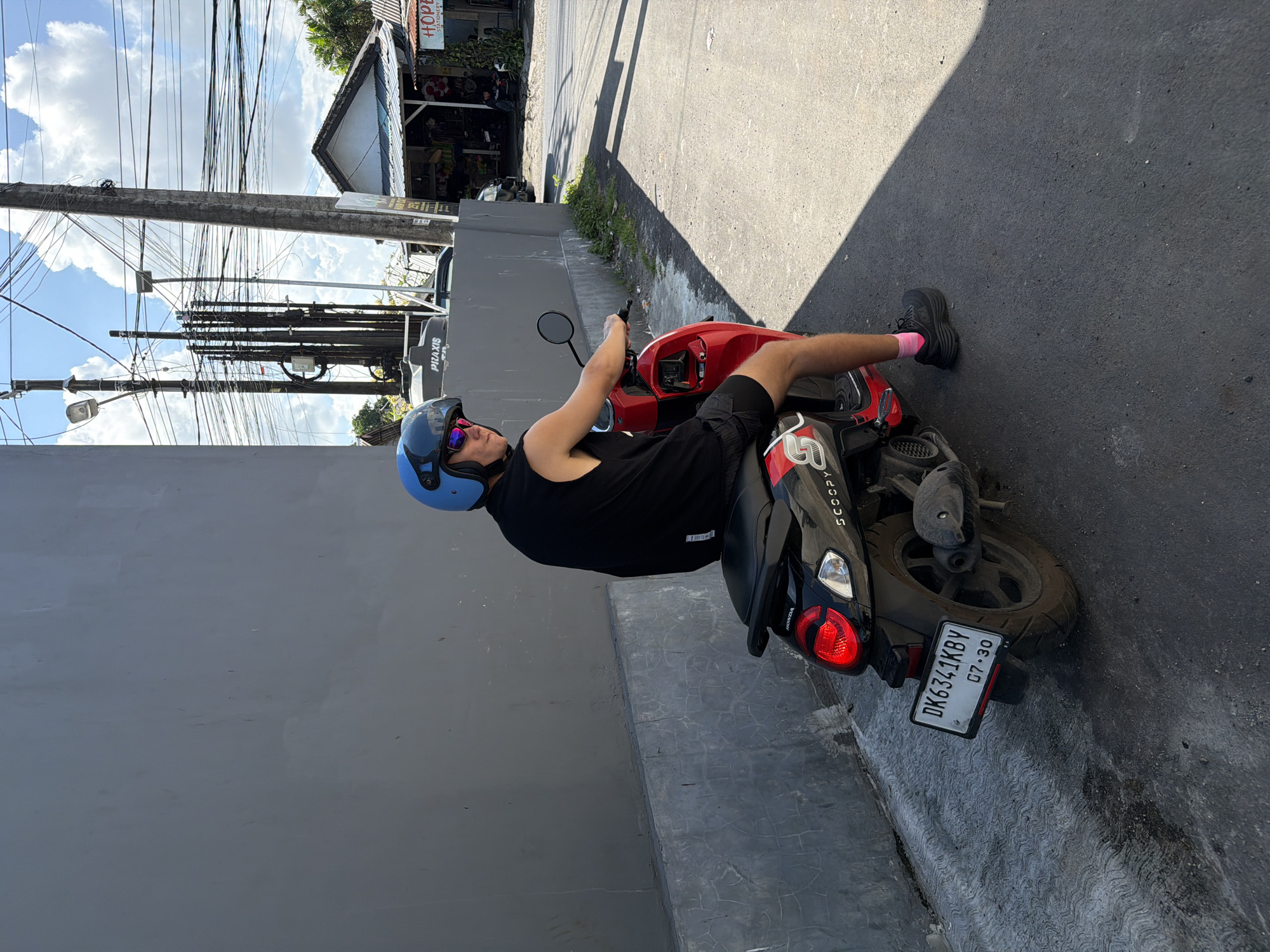 A man is sitting on a red Honda Scoopy scooter on a sunny day in Kuta Selatan, Indonesia. The scooter's license plate reads "DK6341KBY 07-30". The man wears a blue helmet, sunglasses, a black tank top, black shorts, pink socks, and black sneakers. He is holding the scooter's handlebars. The scooter is parked next to a gray wall on an asphalt street. Power lines are visible above. A building with shops and restaurants is in the background.
