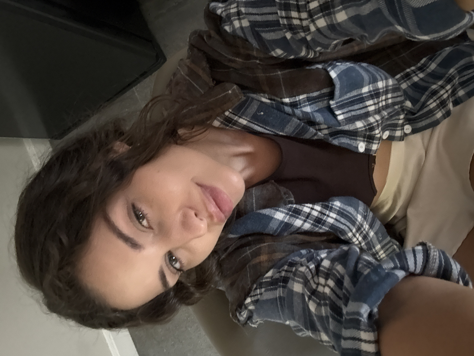 The image is a selfie of a young woman with dark hair and fair skin. She is wearing a brown tank top underneath a blue and brown plaid flannel shirt. The woman's face is close to the camera, showing her eyes, nose, and lips. The background is blurry, but it appears to be an indoor setting. There is no visible indication that the photo was taken in North Kuta, Indonesia.