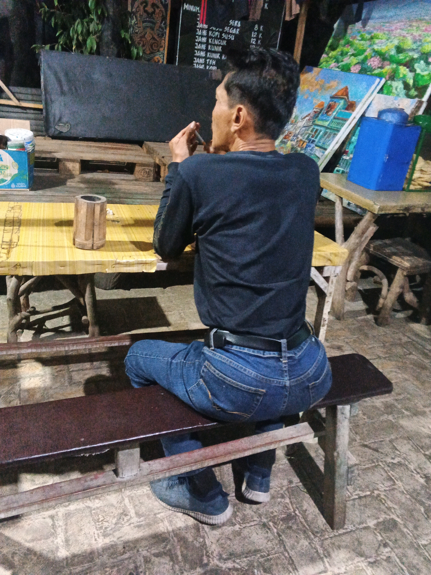 The image depicts a man sitting on a wooden bench in what appears to be an outdoor cafe or stall in Ungaran, Indonesia. The man is wearing a dark long-sleeved shirt and blue jeans. He is facing away from the camera, looking towards a table where he is holding a cigarette in his right hand. On the table, there is a wooden cylindrical object and what looks like a menu board in the background. There are also paintings displayed on easels behind the table. The environment suggests a casual, rustic setting with a cobbled ground surface and wooden furniture. The lighting indicates it might be dusk or evening.