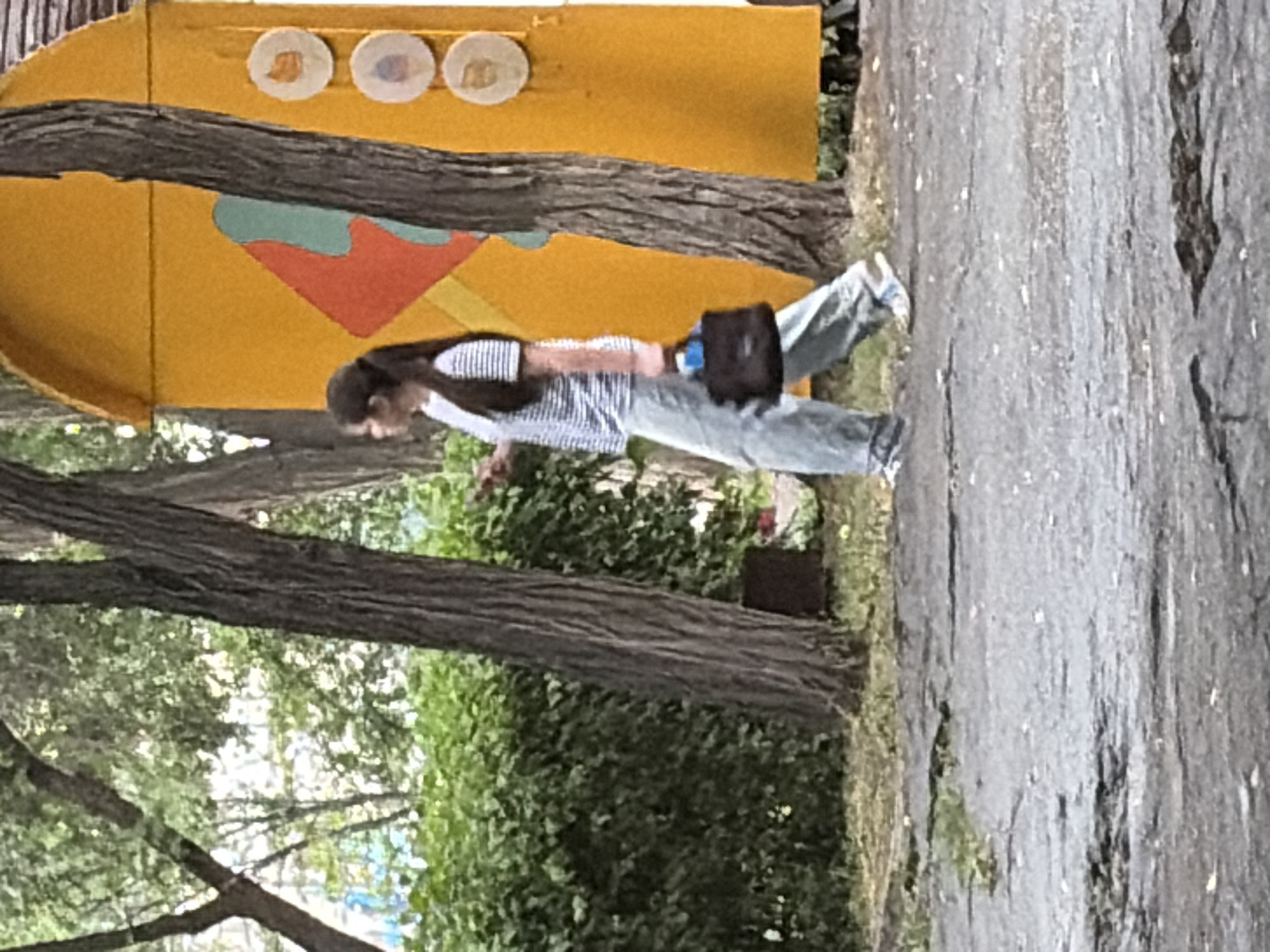 A young woman with long dark hair, wearing a striped t-shirt and light-colored pants, walks to the right, holding a dark handbag. She is walking on a paved path next to a lush green hedge. Behind her, a large, bright yellow structure with colorful geometric designs and circular embellishments stands. The environment appears to be outdoors, possibly a park or a recreational area, with several large trees with rough bark present in the foreground. The ground is a textured, dark asphalt.