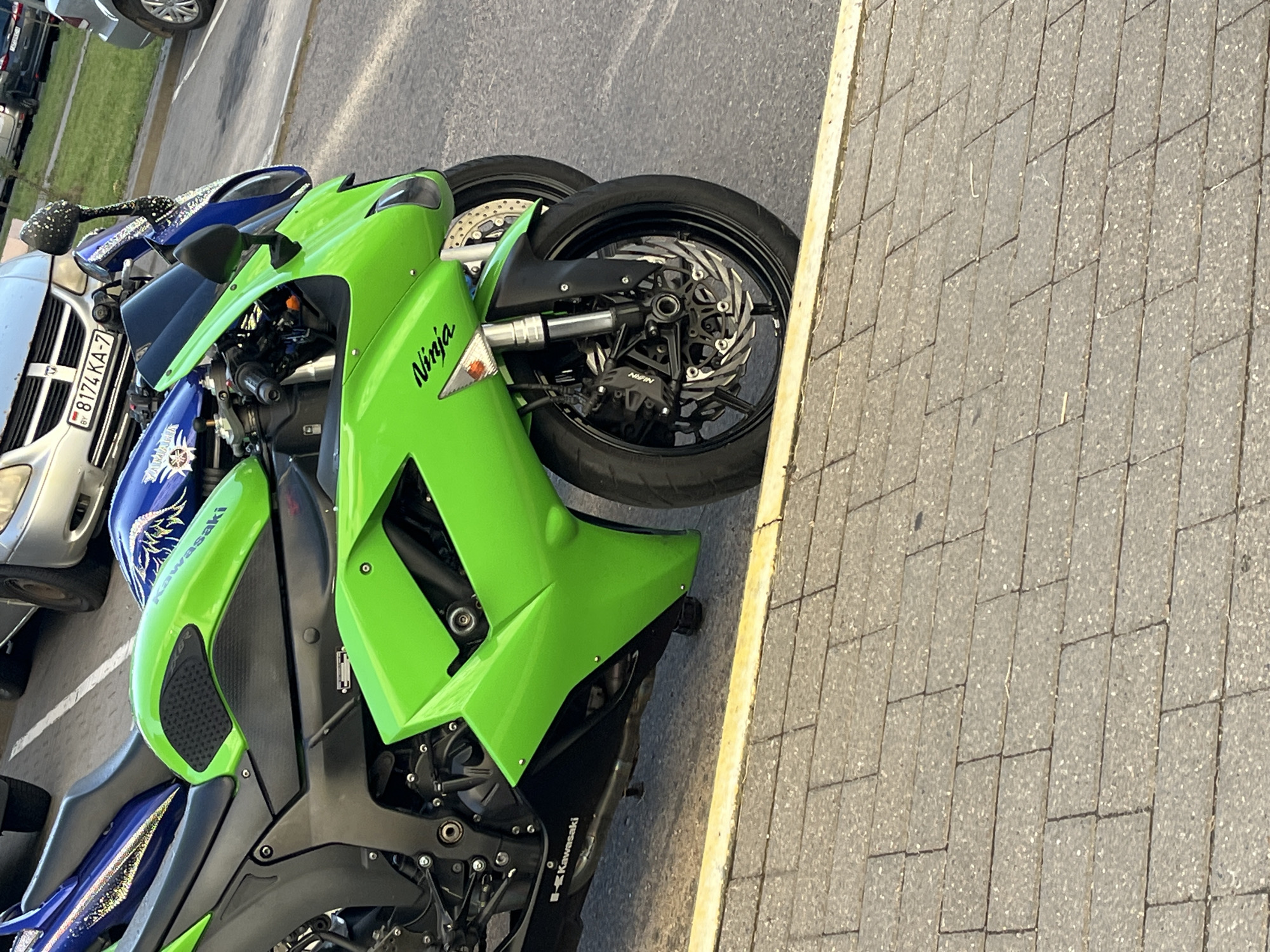 This image shows a close-up view of two motorcycles parked on a street in Minsk, Belarus. The foreground motorcycle is bright green and features "Ninja" branding on its fairing and "Kawasaki" branding on its lower bodywork. It has a black seat and black wheels with prominent front brakes. Behind it, a blue motorcycle is partially visible, also with branding that appears to include "Yamaha". The motorcycles are parked on a paved surface bordering a brick sidewalk. In the background, parts of cars are visible, including a license plate that reads "BY 8174 KA-7". The overall scene suggests a casual parking situation on a city street.