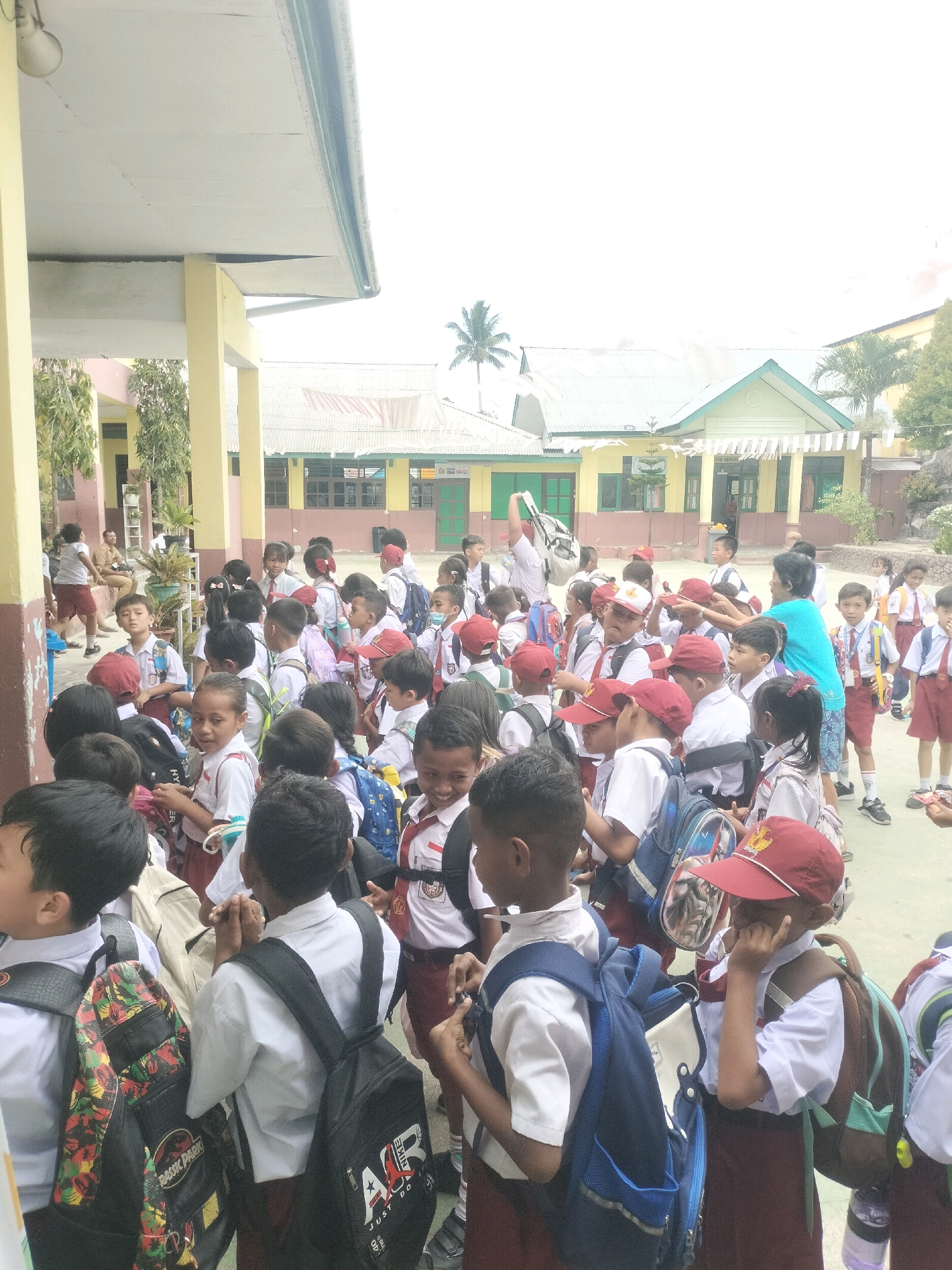 This is a medium-shot, eye-level, full-color photograph of a group of schoolchildren in uniform standing in an outdoor courtyard. The children are wearing white shirts, maroon skirts or shorts, and some have red hats. Most of the children are carrying backpacks. The environment appears to be a school in Dobo, Indonesia, with a covered walkway on the left supported by yellow columns, and a building with a green roof in the background. There are also trees and banners visible. The children are gathered together, with some looking at the camera and others interacting with each other. The overall scene suggests a typical school day activity, possibly arriving at school or waiting for a class to begin. The lighting is bright, indicating it is daytime, and the sky is overcast.