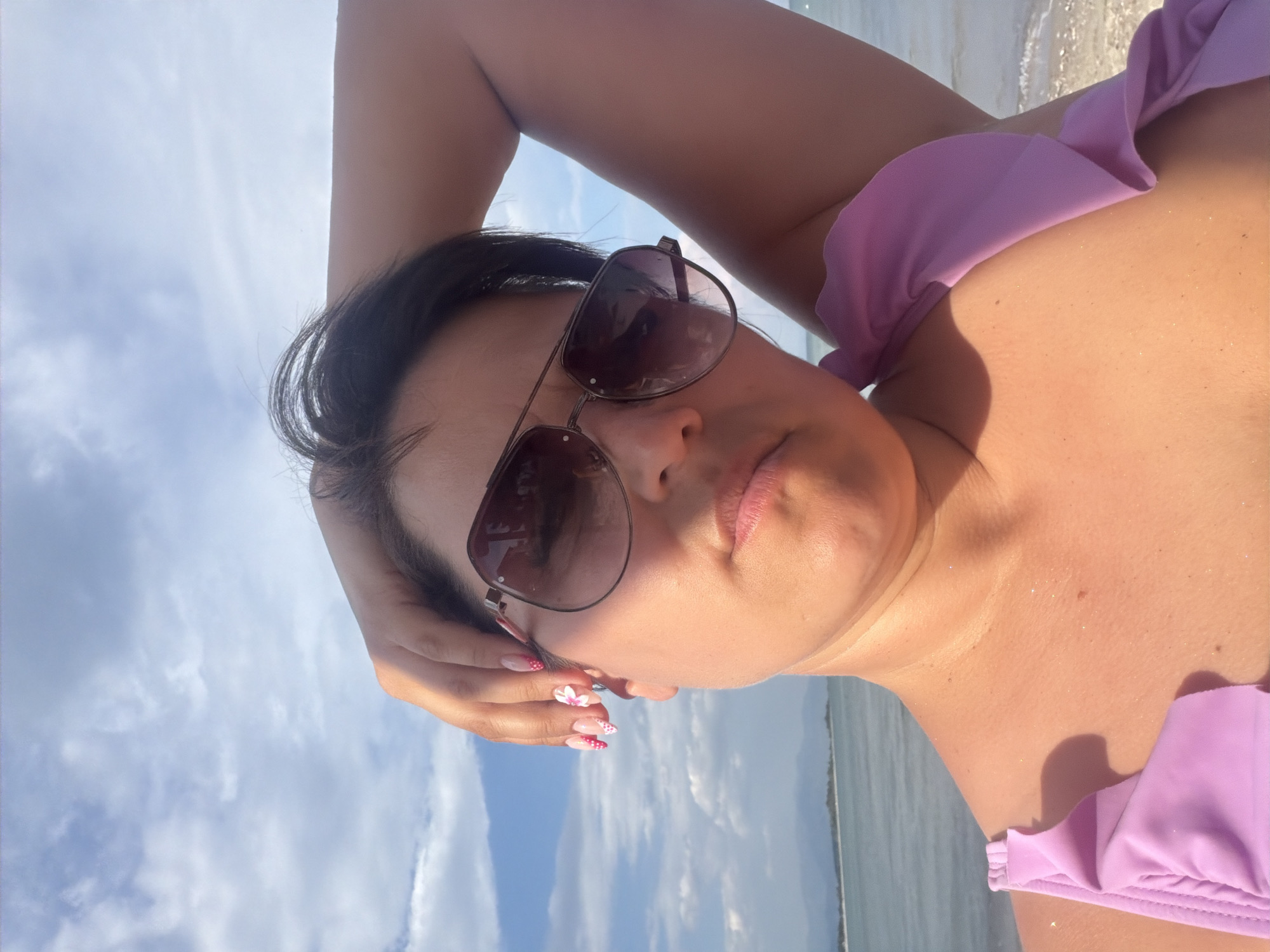 This media file is a close-up, angled selfie taken by a person at a beach. The subject is a woman wearing sunglasses and a lavender bikini top. Her arm is raised behind her head, and her hand is resting on her hair. Her nails are painted with a pink and white polka dot design and small flower embellishments. The background shows a bright sky with scattered clouds and the ocean with gentle waves. The lighting suggests it is daytime, likely sunny, and the overall scene conveys a relaxed, vacation atmosphere. The woman's facial expression is neutral, and she appears to be enjoying a leisurely day by the sea. There is no visible text in the image.