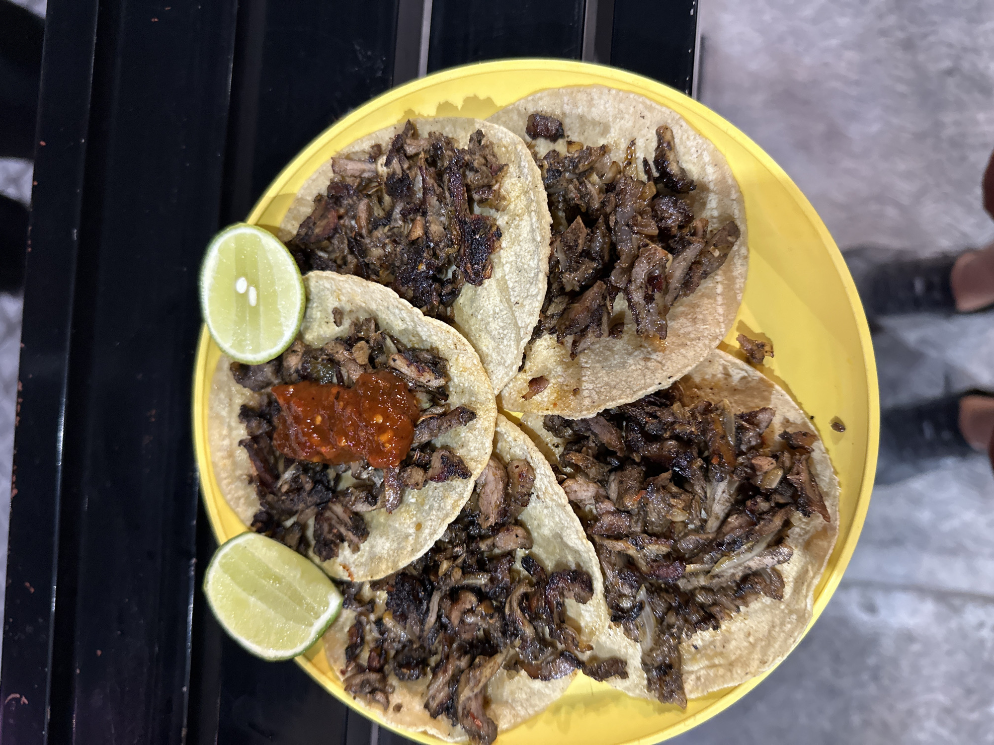 A bright yellow circular plate, positioned centrally in the image, holds six tacos. Each taco consists of a small, light-colored corn tortilla generously filled with dark, finely chopped meat, exhibiting a seared or grilled appearance with some darker, possibly charred, fragments. One taco, located near the top-center, is topped with a distinct dollop of reddish-orange, chunky salsa. Two lime wedges, cut horizontally, are placed on the plate: one in the upper left quadrant and another in the upper right quadrant. The plate rests on a dark, horizontally ridged surface, likely a table or counter. Below the plate, a light gray paved surface is visible, along with partial views of dark footwear, indicating a person standing over the scene. This scene is situated in San Bernardino Tlaxcalancingo, Mexico.