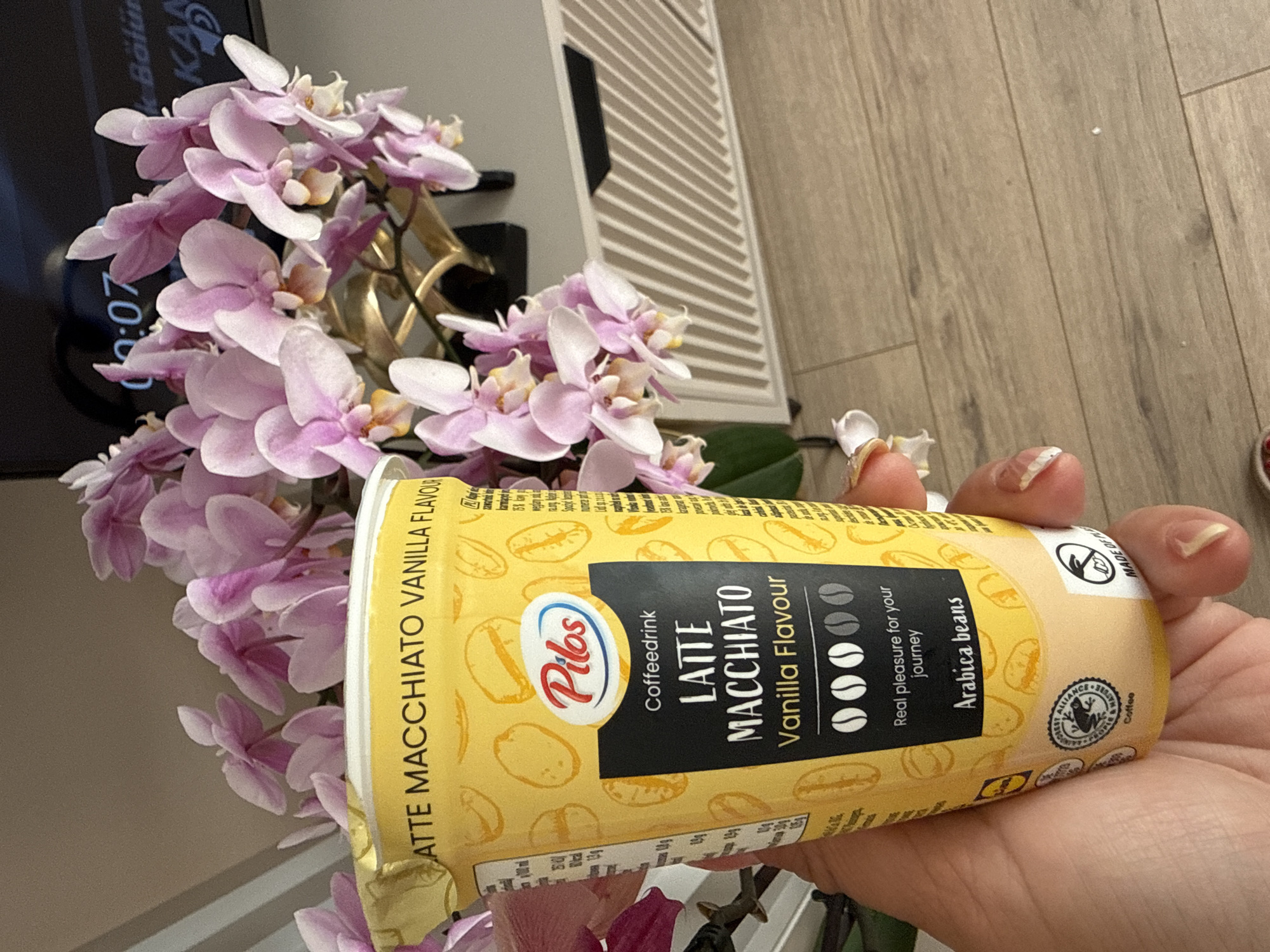 A hand holds a yellow cup of Pilos brand coffee drink with a vanilla flavor. The cup has a picture of coffee beans and the words "LATTE MACCHIATO Vanilla Flavour" on it. Behind the cup, a bouquet of pink orchids is visible, and a portion of a television screen shows a clock reading "00:07" and text in Turkish. The scene appears to be set indoors, with a wooden floor and a light-colored cabinet in the background.