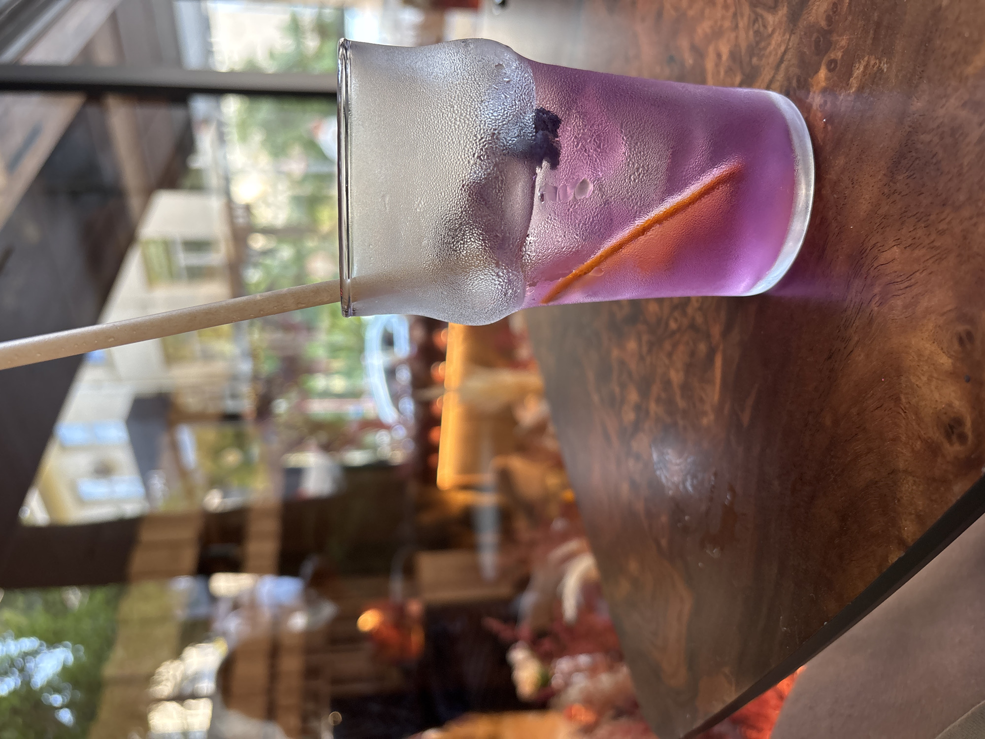 A frosted glass with a purple liquid and a straw rests on a dark wood table. The glass contains ice cubes, a slice of orange, and a dark berry. The background is blurred, showing reflections of buildings and greenery, suggesting an outdoor seating area of a cafe or restaurant. The overall scene evokes a refreshing and relaxed atmosphere.