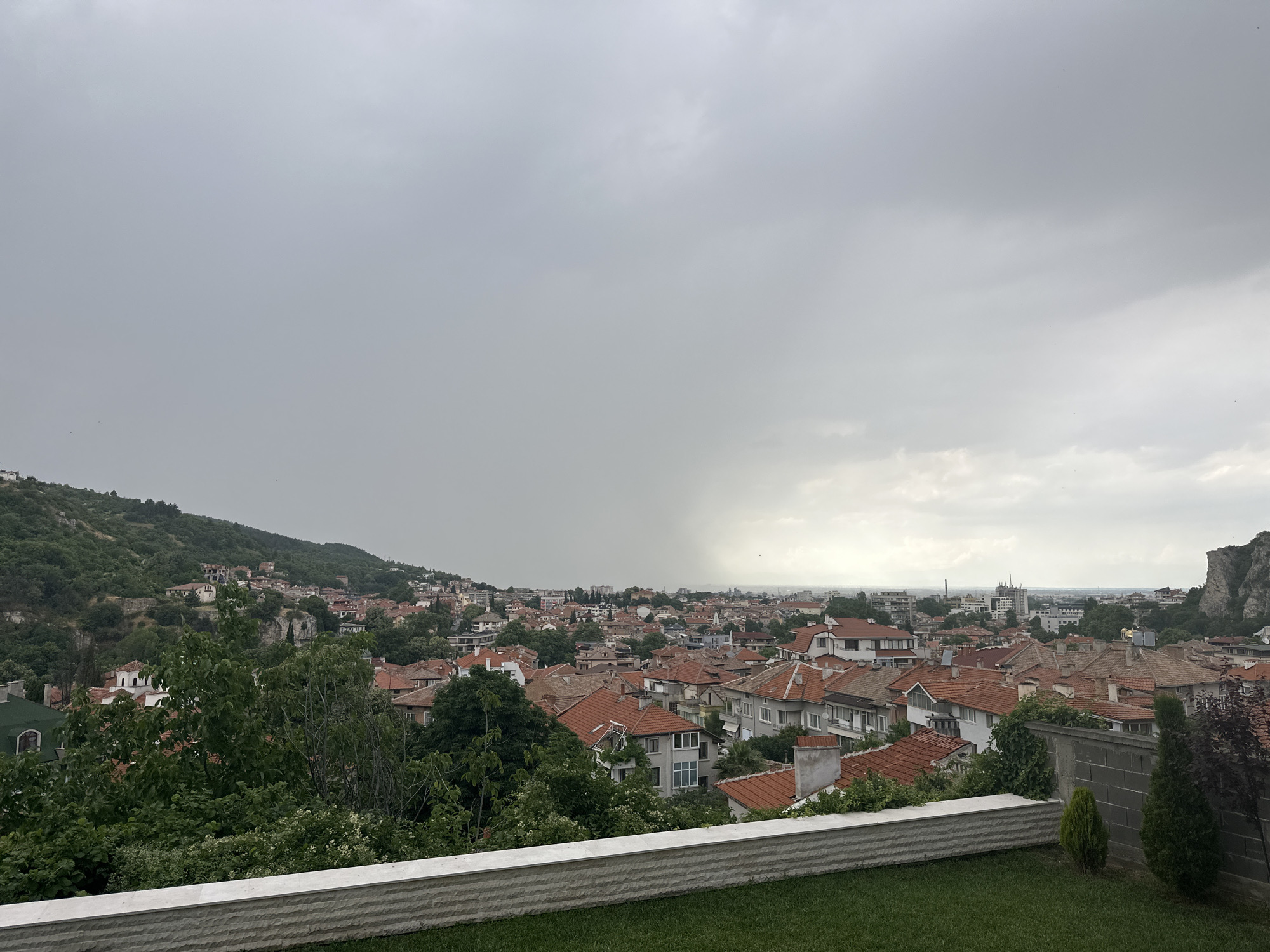 This image depicts a panoramic view of Asenovgrad, Bulgaria, from an elevated vantage point. The foreground features a meticulously maintained lawn and a low, white, stone-patterned wall. Beyond the wall, dense green trees and foliage dominate the lower half of the frame, obscuring some of the buildings.
The middle ground is filled with a dense cityscape characterized by numerous residential buildings with red-tiled roofs, a common architectural style in the region. Some of these buildings are modern, while others appear older. The city stretches towards the horizon, with a mix of urban development and natural landscape. In the background, a lush, green, tree-covered hillside rises, dotted with more structures and ancient-looking buildings, possibly part of the historical city. To the far right, a dramatic rock formation with steep cliffs juts out above the cityscape. The sky is overcast and gray, suggesting a cloudy or potentially rainy day, with a faint, brighter area on the horizon possibly indicating the direction of the sun behind the clouds. No people or animals are visible in the frame.