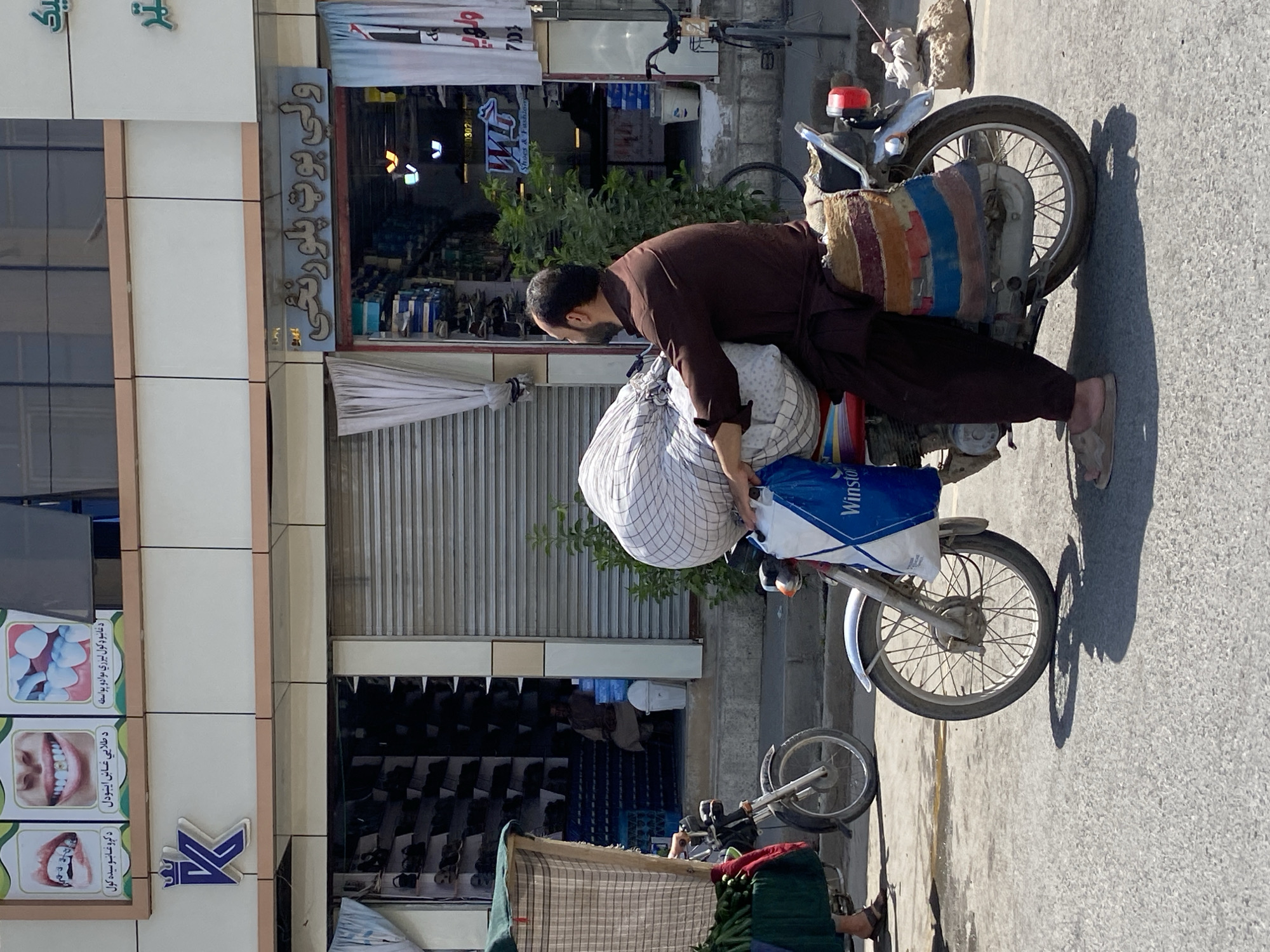 A man wearing a brown shalwar kameez is riding a motorcycle laden with a large, checkered bundle and a blue plastic bag. He is wearing sandals. The motorcycle has a colorful textile bag attached to the rear fender. The scene appears to be in Kandahar, Afghanistan, indicated by the Dari script on the storefronts and the architecture. Behind the man, a storefront displays shoes and fashion items, with a roller shutter partially closed. Another motorcycle, partially visible on the left, is also loaded with produce. The overall environment is an outdoor marketplace or street with shops and buildings lining the road.
