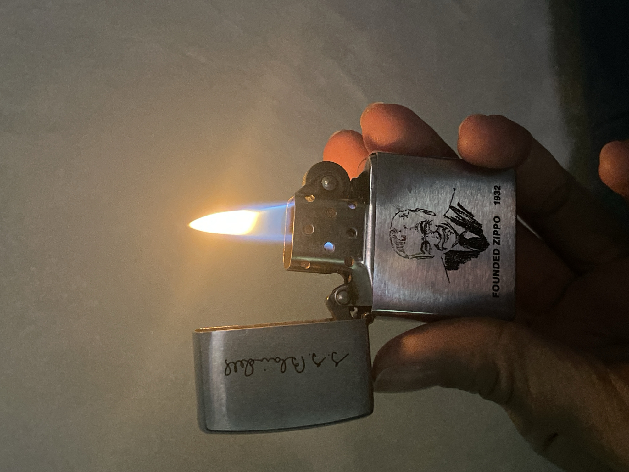 A hand holds a Zippo lighter with a lit flame. The lighter is silver and has an image and the words "FOUNDED ZIPPO 1932" printed on it. The inside of the lid has writing on it. The background is blurry and light-colored. There is no indication of the location being Izhevsk, Russia.