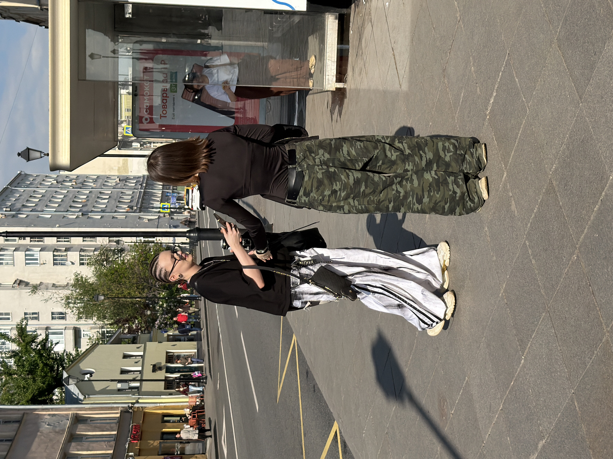 The image captures a street scene in Moscow, Russia, showing two individuals standing on a sidewalk next to a road. The person closer to the camera has braided hair, sunglasses, and wears a black top paired with white and black patterned pants. They carry a small black bag with a chain strap. The other person wears a black top and camouflage print pants. They are standing in front of a storefront with reflections visible in the glass. Buildings are visible in the background, and the road has yellow lane markings. The scene is well-lit, suggesting it is daytime.
