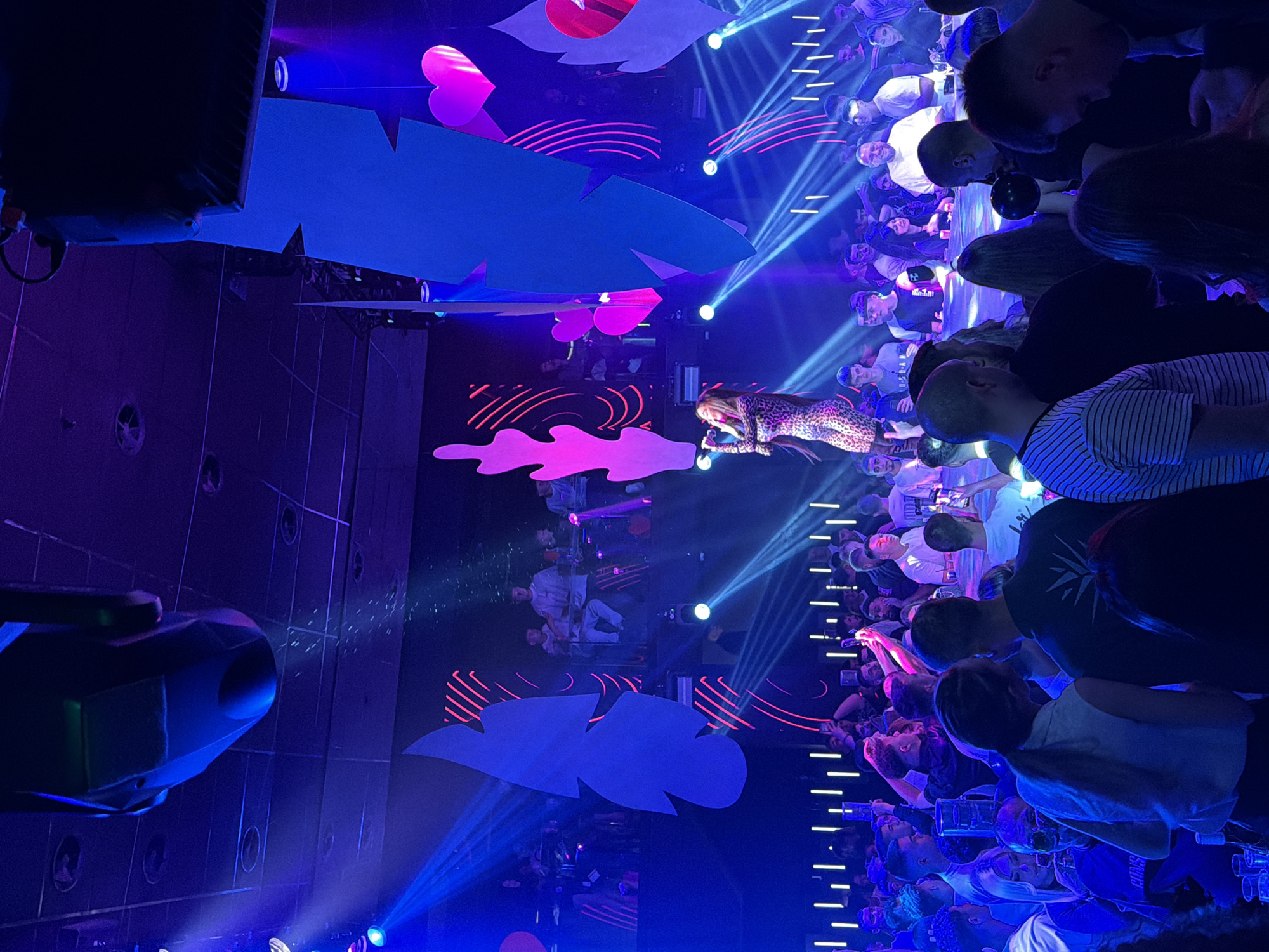 A crowded indoor venue, likely a concert hall, is filled with concertgoers watching a female performer on stage. The performer, wearing a patterned bodysuit, is singing into a microphone. The stage is decorated with large cloud and heart-shaped props. Blue stage lighting illuminates the scene. Laser beams cut through the crowd, adding to the atmosphere. The audience members are a mix of genders and ages. They're holding their phones up, recording the performance. The venue location is Blagoevgrad, Bulgaria.