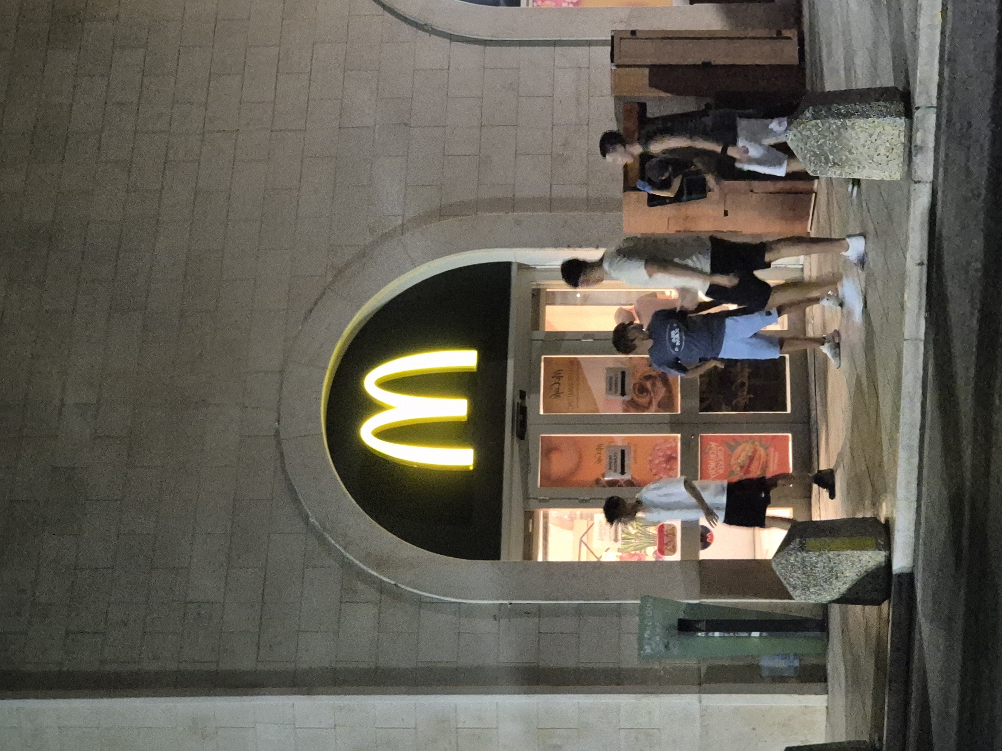The image shows the exterior of a McDonald's restaurant at night, located in the city of Taranto, Italy. A large, illuminated golden arch logo is visible above the entrance. Several young people, appearing to be teenagers or in their early twenties, are standing outside the restaurant. Some are facing the entrance, while others are interacting with each other. The ground in front of the building is paved, and there are several concrete bollards. The architecture of the building features light-colored stone blocks. The lighting suggests it is evening or nighttime.
