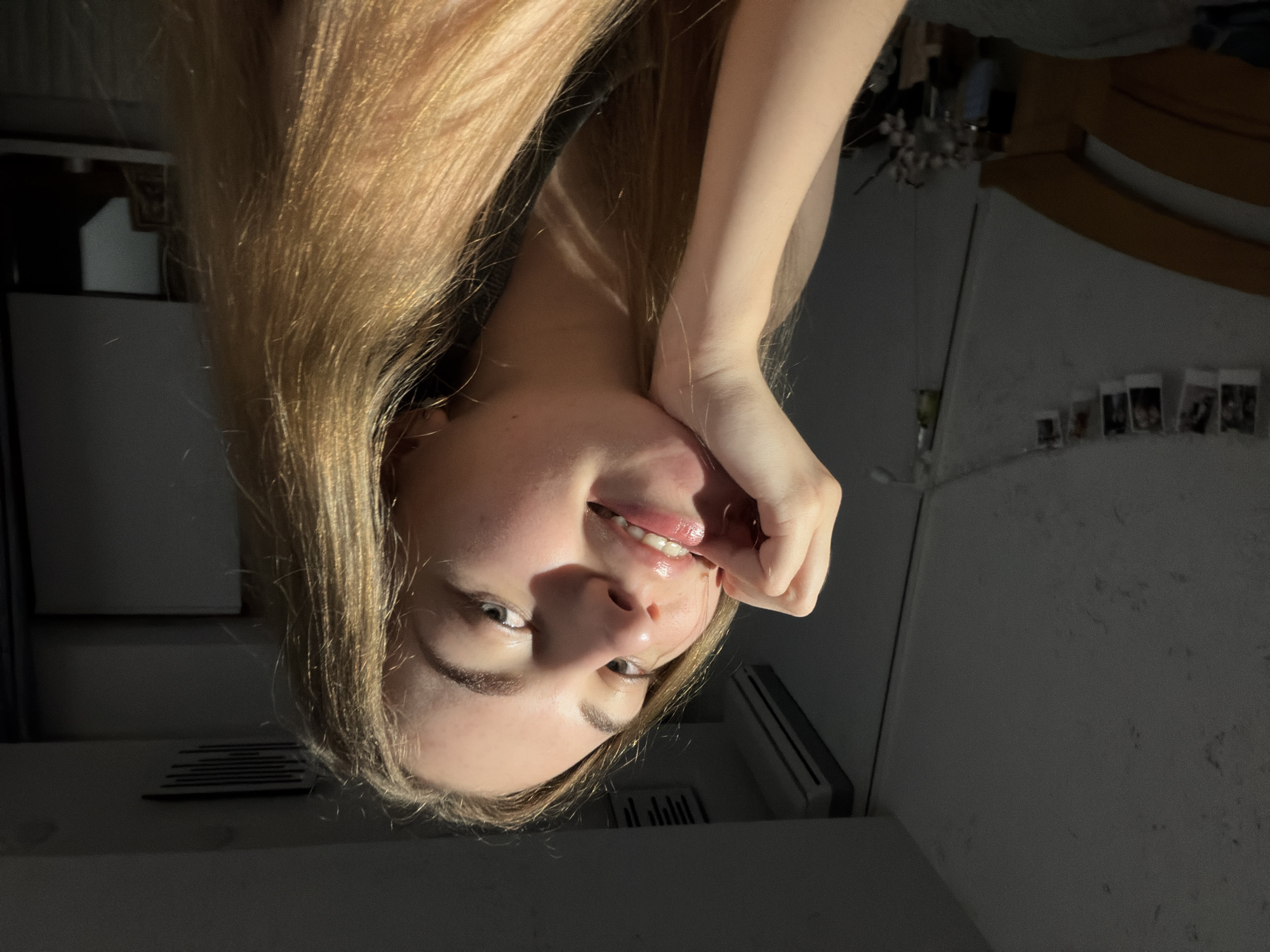 A young woman with blonde hair is photographed from the chest up, looking towards the camera with her hand resting on her chin. She is in a dimly lit room, with a window casting a strong light on her face, creating a shadow on the left side of the image. The background is mostly dark, with a hint of a bed frame on the left and what appears to be a mirror and a picture frame on the right. There are also some photos hanging on a string in the background. The overall impression is that of a casual, indoor portrait taken in a residential setting. The provided crops do not offer any specific details about the location within the city of Dnipro, Ukraine.
