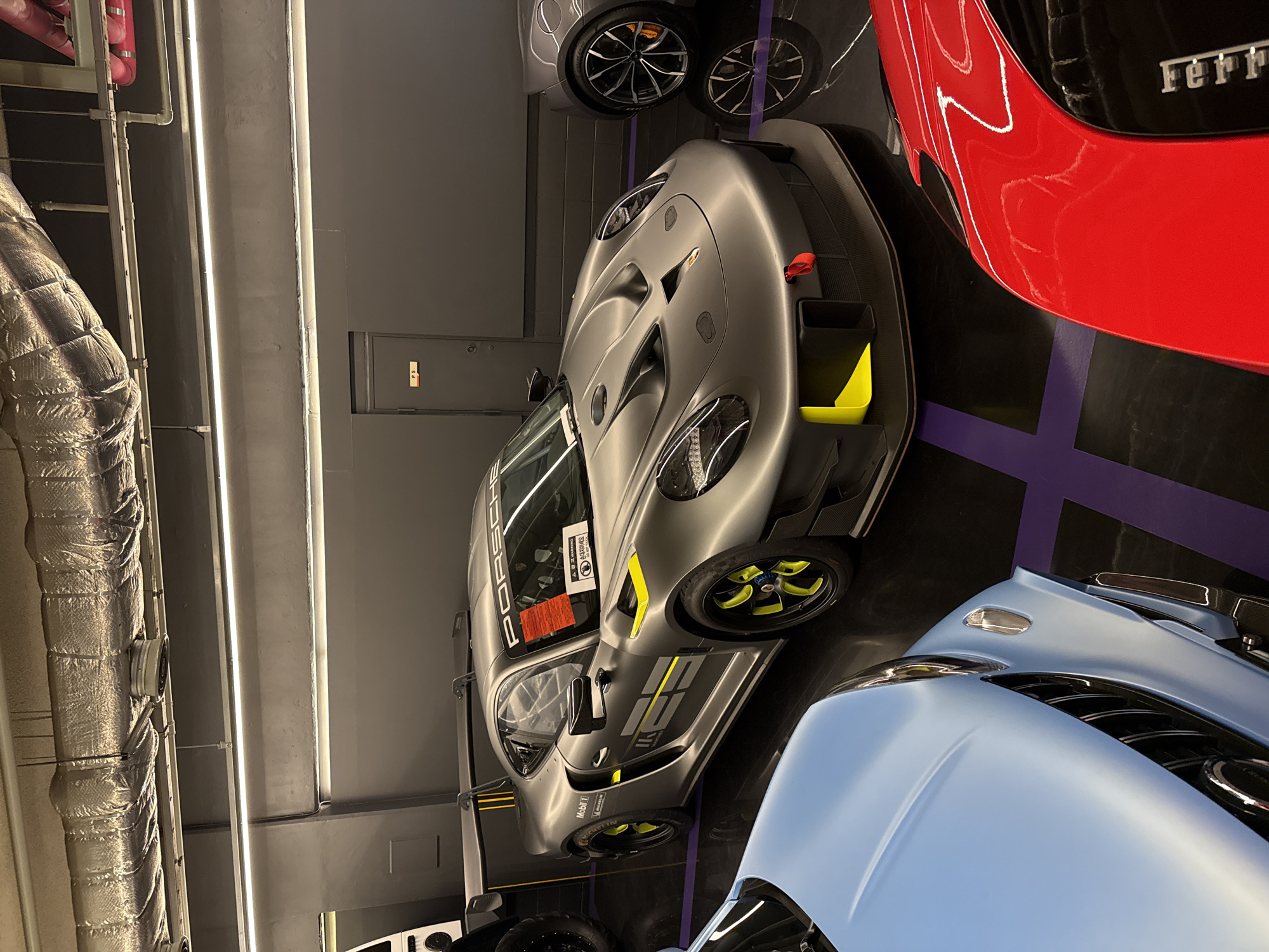 The image depicts an indoor showroom containing multiple high-end sports cars. A grey Porsche race car with neon yellow accents is centrally positioned. Beside it are parts of a red Ferrari and a light blue sports car. The floor is marked with purple tape. The ceiling contains industrial features like pipes and strip lighting. The setting appears to be a car dealership in an urban environment. No people are directly visible in the shot. The location in Seoul, South Korea is not apparent in the picture.