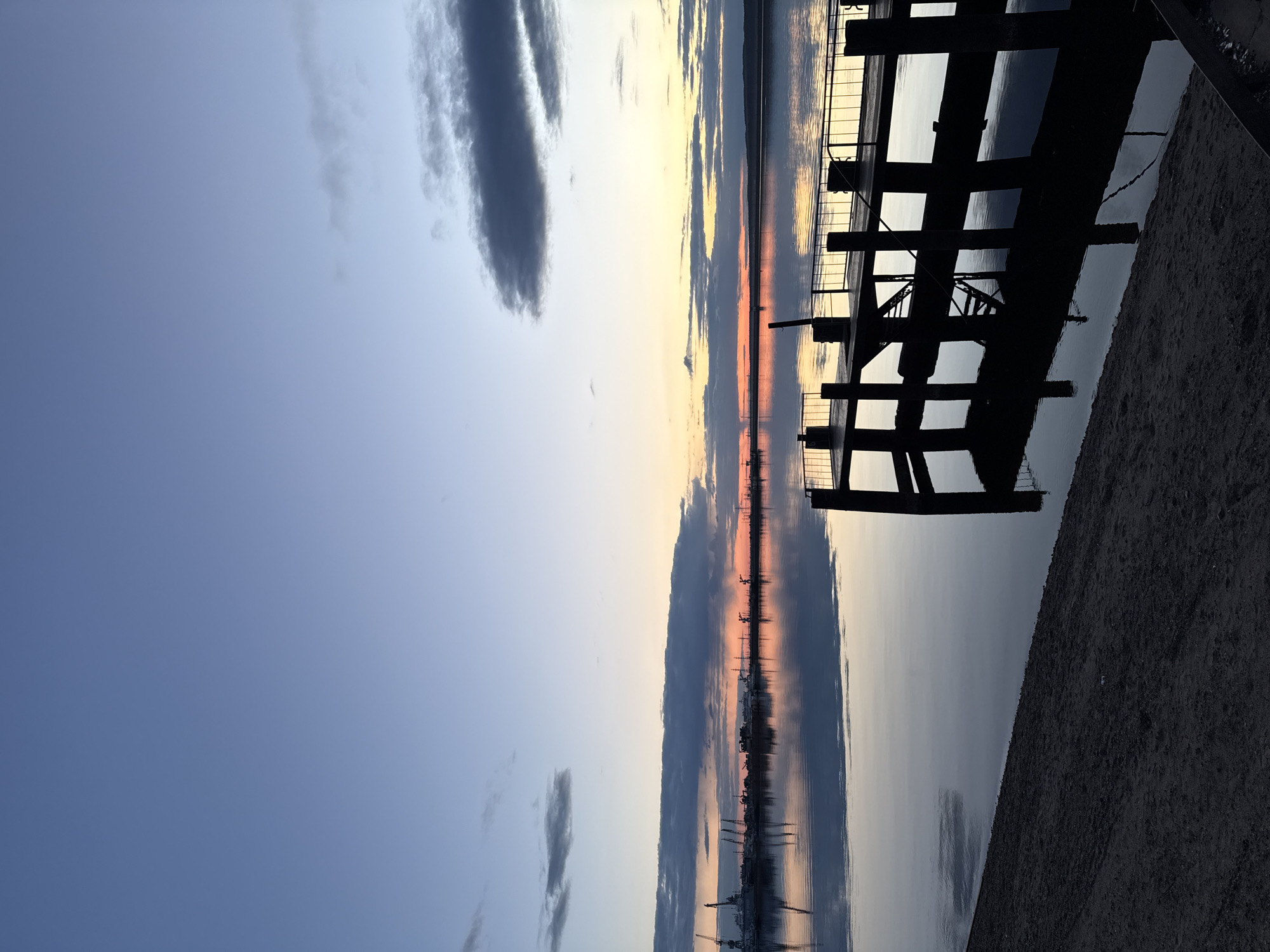This landscape image depicts a tranquil scene at either sunrise or sunset over a large body of water, consistent with the Votkinsk Reservoir near Chaikovsky, Russia. The sky, occupying the upper portion of the frame, transitions from a soft blue on the left to lighter blues, yellows, and oranges near the horizon, indicating the low angle of the sun. Several elongated, dark gray clouds are scattered across the sky, with one prominent cloud stretching horizontally in the upper left.
The expansive water surface is remarkably calm and acts as a mirror, perfectly reflecting the colors of the sky and the forms of the clouds. Along the distant horizon, faint silhouettes of what appear to be industrial structures or cranes are visible, their reflections clearly rendered in the still water, particularly where the sky glows with warm orange hues.
In the right foreground, a dark, silhouetted pier or dock structure extends into the water. Its structural elements, possibly beams and railings, create a strong, angular contrast against the soft backdrop of the sky and water. A narrow strip of dark, sandy or gravelly ground forms the extreme right edge of the image, leading to the base of the pier. No people or visible text are present. The overall impression is one of peaceful natural beauty, enhanced by the reflective quality of the water and the dramatic sky colors.
