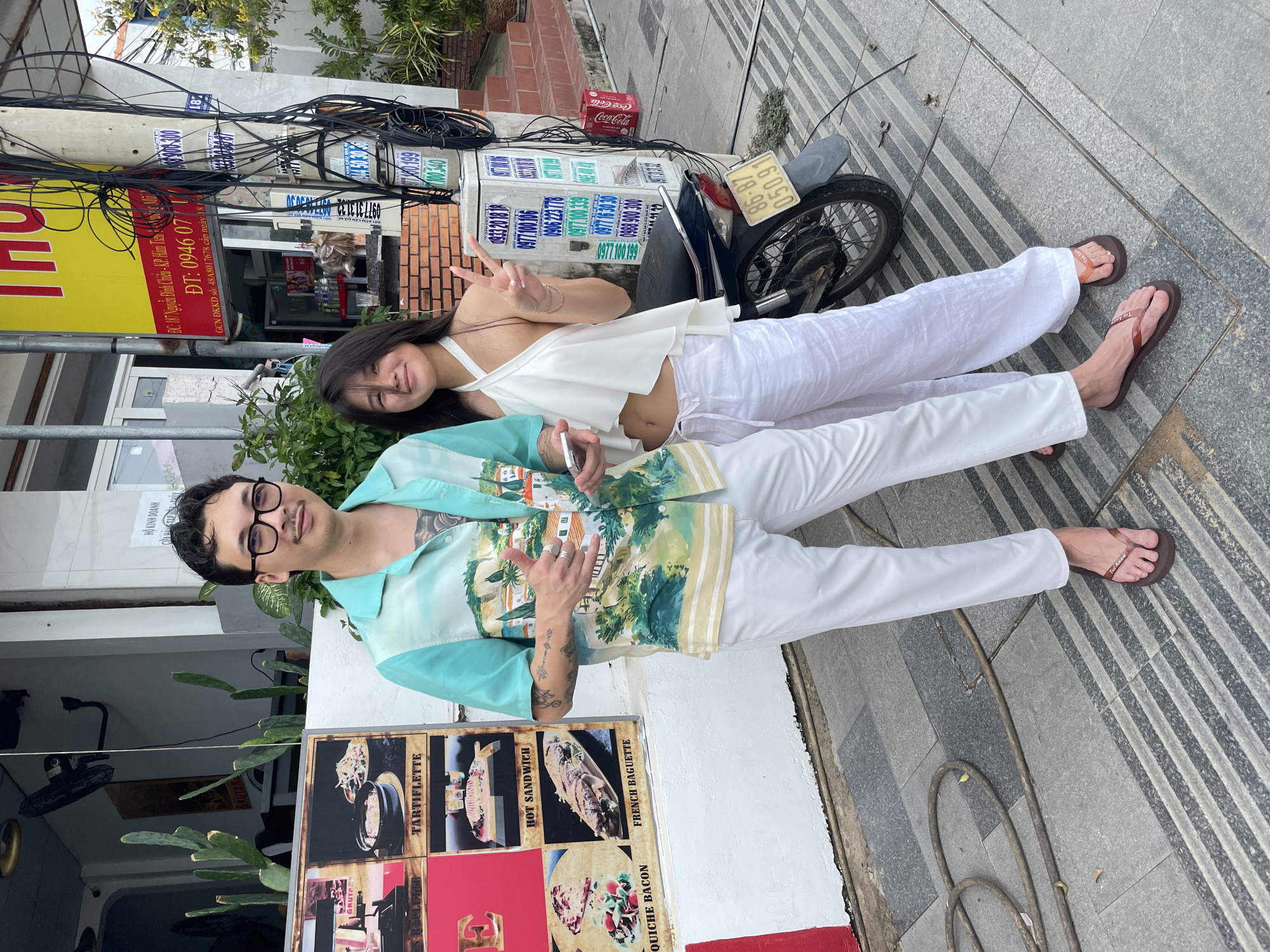 **Scene:**
A photo taken in what appears to be the Phu Thuy Ward in Vietnam.
**People:**
A woman and a man are posing for the photo. The woman has long, dark hair and is wearing a white top and pants. She is holding up a peace sign. The man has short, dark hair, glasses, and a mustache. He is wearing a short-sleeved shirt with a patterned design and white pants. He is also making a gesture with his hands.
**Objects:**
A motorbike is parked behind the woman. There is a Coca-Cola box and a sign with Vietnamese writing on it. A menu displaying food items with names such as "TARTIFLETTE," "HOT SANDWICH," "FRENCH BAGUETTE," and "QUICHE BACON" is also visible. Various electric cables are overhead.
**Environment:**
The scene appears to be on a city street with a building in the background. Sidewalk is striped.